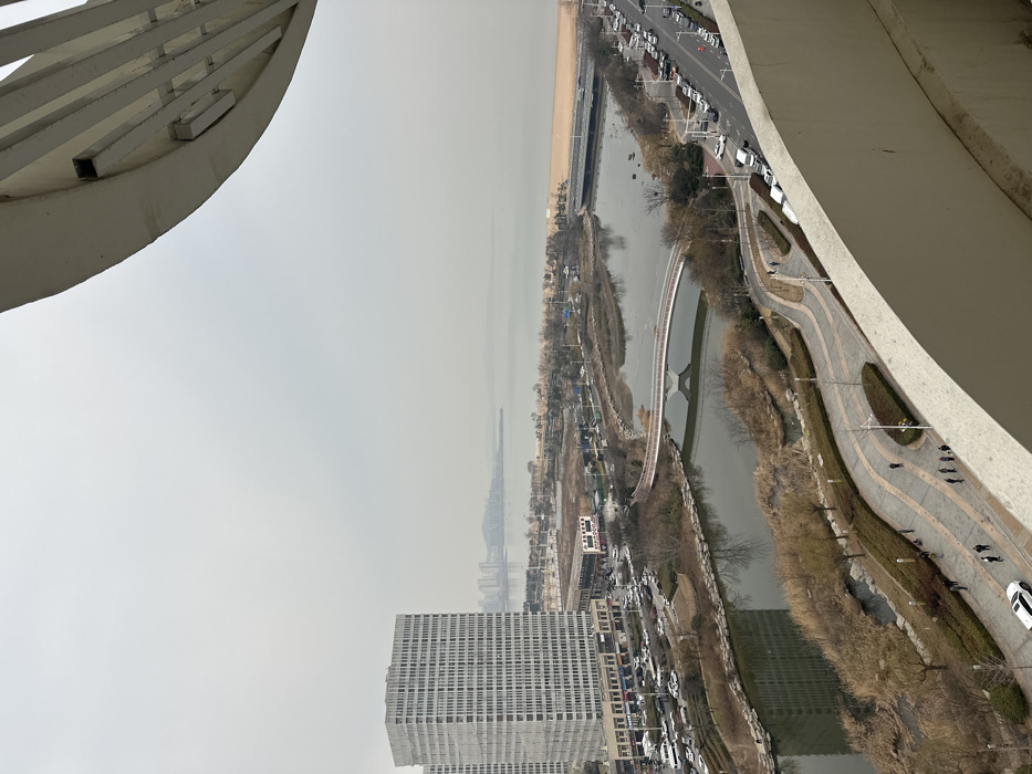 An elevated, wide-angle view captures a segment of Huangdao District, China, under a uniformly overcast and hazy sky. The immediate foreground is partially obscured by a curved, beige concrete architectural element, suggesting a vantage point from a balcony or high-rise.
A winding river or canal, reflecting the muted sky, traverses the midground. On the left bank, a prominent multi-story, white rectangular building, distinguished by a grid of windows, dominates an urban block with several smaller structures and parking areas containing numerous vehicles. Opposite, on the right bank, a multi-lane road parallels the waterway, densely lined with parked cars and a few moving vehicles.
Two bridges span the river: a white, arched pedestrian bridge is visible in the mid-left, and a wider, darker vehicular bridge is positioned further downstream towards the background. Beyond these bridges, the river opens into a large body of water, likely a bay or the sea, with a vast sandy beach bordering its near shore. A distant, hazy cityscape is discernible across the water on the horizon.
Along the riverbanks, particularly on the right, landscaped pedestrian pathways with undulating designs are present. These paths are flanked by bare deciduous trees, shrubs, and brown vegetation, suggesting a late autumn or winter season. Multiple streetlights line the pathways. Several individuals, appearing as small, dark figures, are observed walking on these pedestrian routes.