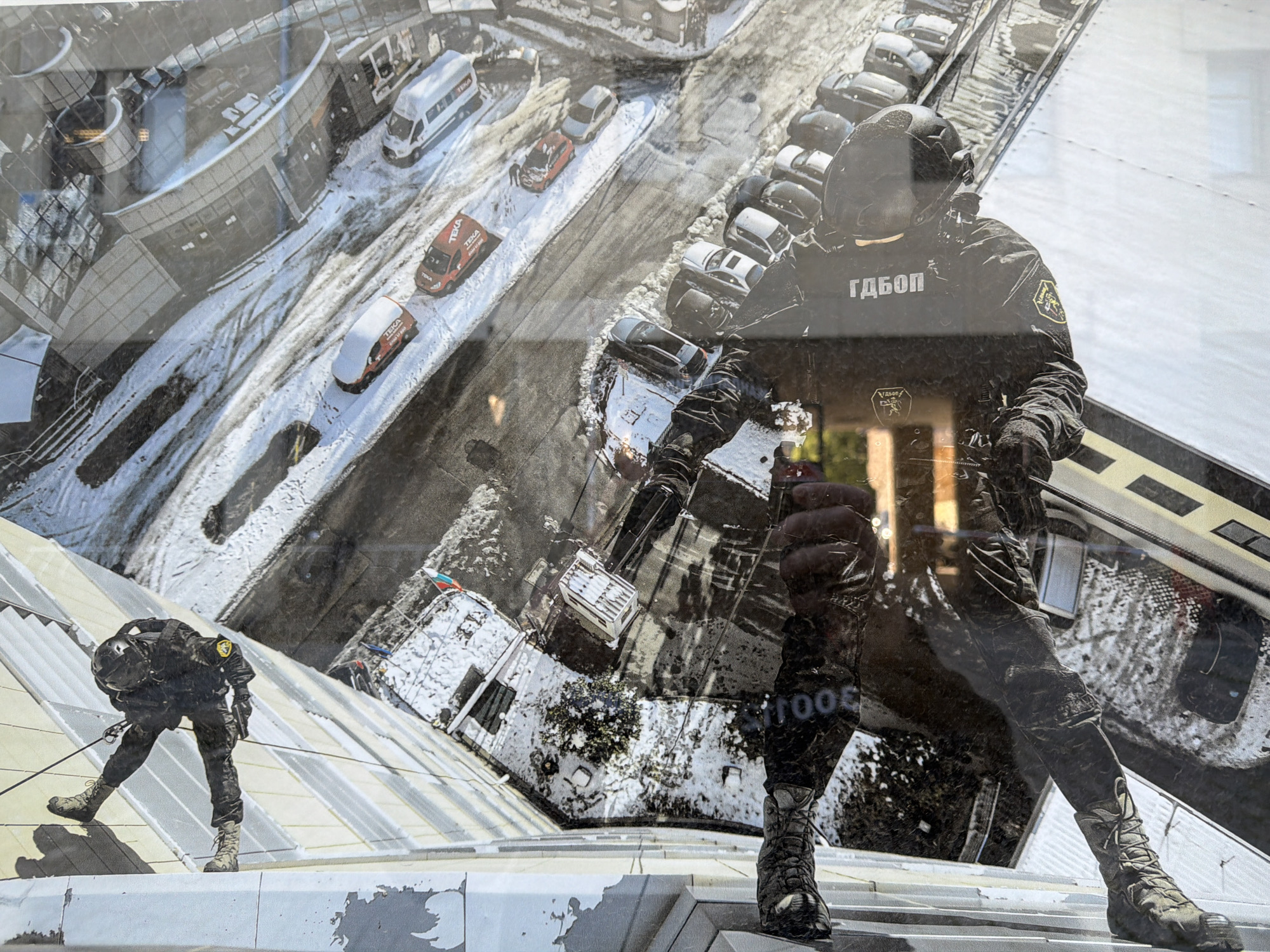 The image depicts two armed men rappelling down the side of a building in Sofia, Bulgaria. One man is visible in the lower left, fully equipped, rappelling down a snow-covered slanted rooftop. The other is positioned higher, partially obscured by a reflection in the window. The reflection shows another armed man, and parts of an urban scene with cars, buildings and snow. "ГДБОП" is written on the armed men's uniforms.