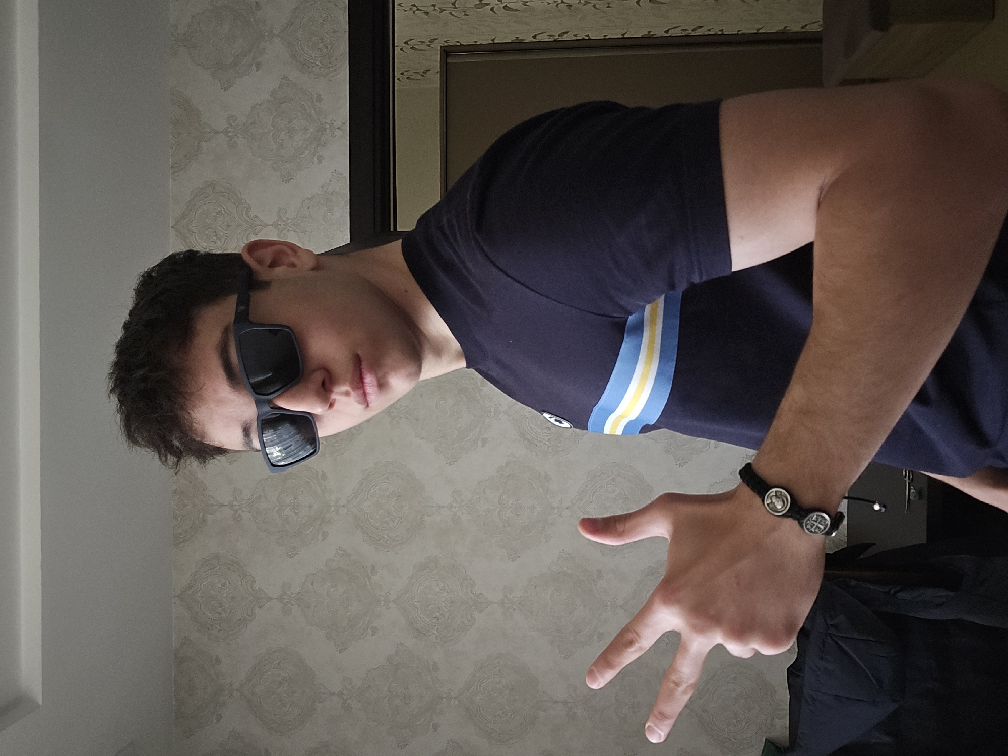 A young man wearing sunglasses and a dark blue T-shirt with yellow and white stripes on the sleeve is posing indoors. He has short, dark curly hair and is making a hand gesture. He wears a black bracelet with silver medallions. The background shows a patterned wallpaper and a partial view of a dark-framed mirror. The location cannot be confirmed as Yerevan, Armenia without additional context.