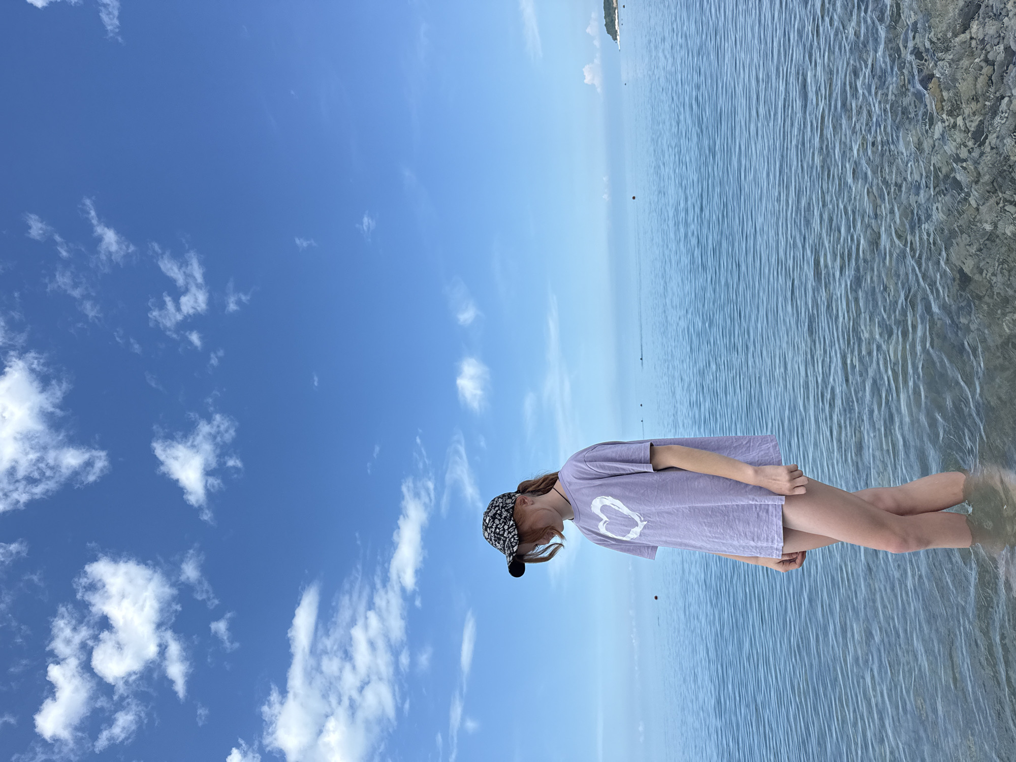 A young person with red hair, wearing a patterned baseball cap and a light purple t-shirt with a white heart design, stands in shallow, clear water. They are looking down. The scene is a body of calm blue water, likely the Black Sea, with a clear blue sky dotted with white clouds above. In the distance, a small island or landmass with trees is visible on the right horizon. The water has ripples and a rocky bottom is visible near the foreground. This location is consistent with the coastline of Nebug, Russia.
