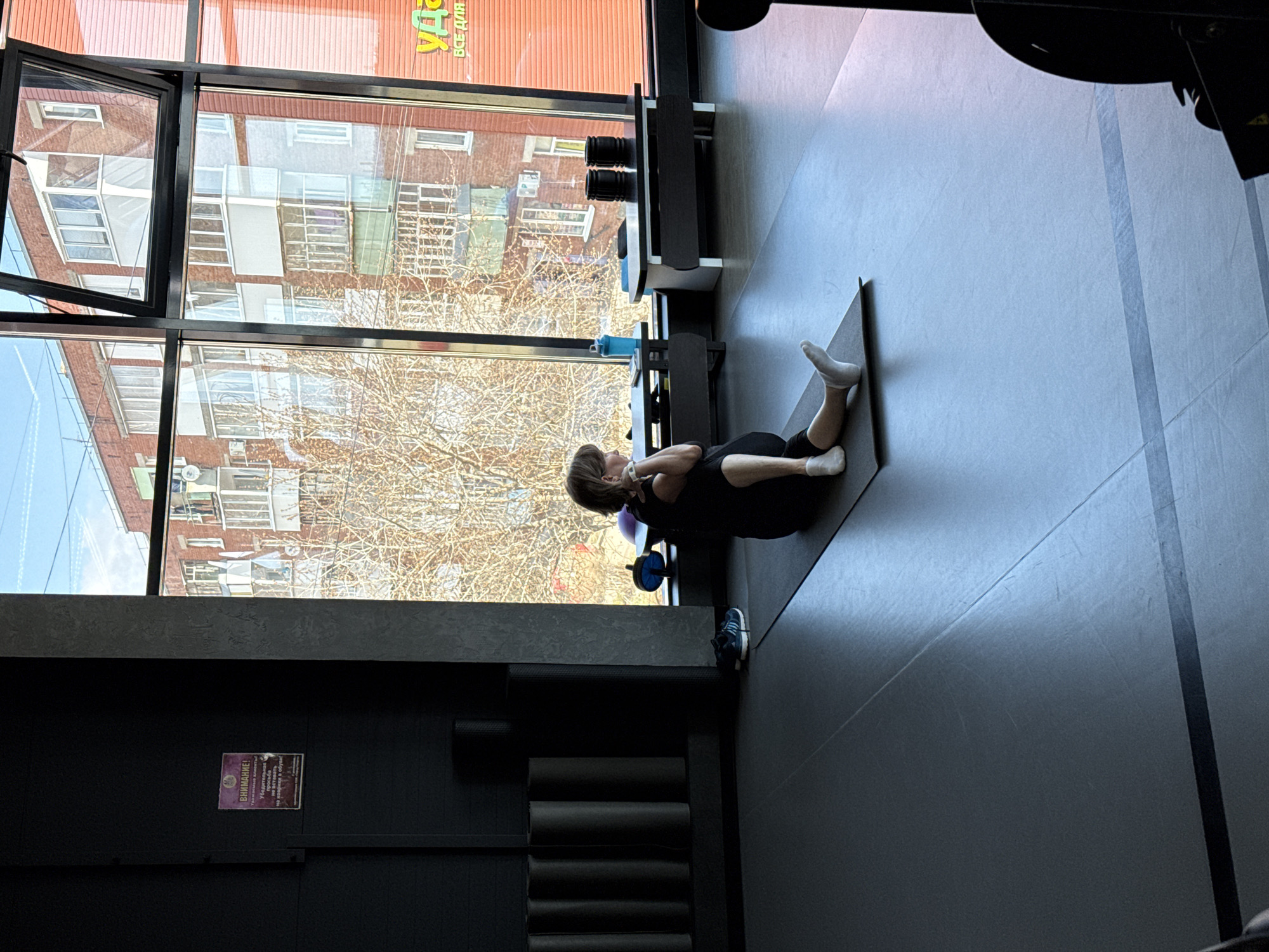 This media file captures an indoor scene within a modern gym or fitness studio, overlooking an urban landscape. The primary subject is a person, likely female, lying on their back on a dark exercise mat and performing an abdominal exercise, possibly a crunch or sit-up. They are wearing a dark athletic top and shorts, with white socks on their feet. A small purple ball or cushion is placed under their neck.
The setting features large, black-framed windows that dominate the background, offering a view of multi-story residential buildings with balconies, typical of an urban environment in Votkinsk, Russia. The trees outside have bare branches, suggesting it is late autumn, winter, or early spring. Bright daylight streams through the windows, illuminating the scene.
Inside the studio, the decor is minimalist with dark, possibly black, walls and floor. To the left of the windows, a shelf holds several rolled-up black exercise mats. Near the person exercising, another piece of equipment, possibly a dumbbell or weight, is visible next to the window. A pair of blue athletic shoes rests on the floor nearby. On a black wall, a purple sign with white Cyrillic text is visible. It reads "ВНИМАНИЕ!" (ATTENTION!) at the top, followed by "Убедительная просьба..." (Urgent request...), likely detailing gym rules or instructions. Through the windows, on a building across the street, part of a commercial sign in Cyrillic is visible, with green text reading "Все для" (Everything for) and red text below it, possibly "ТОВАРЫ для" (Goods for...).
The overall impression is a focused workout session in a well-lit, contemporary fitness space with a clear view of the city outside.