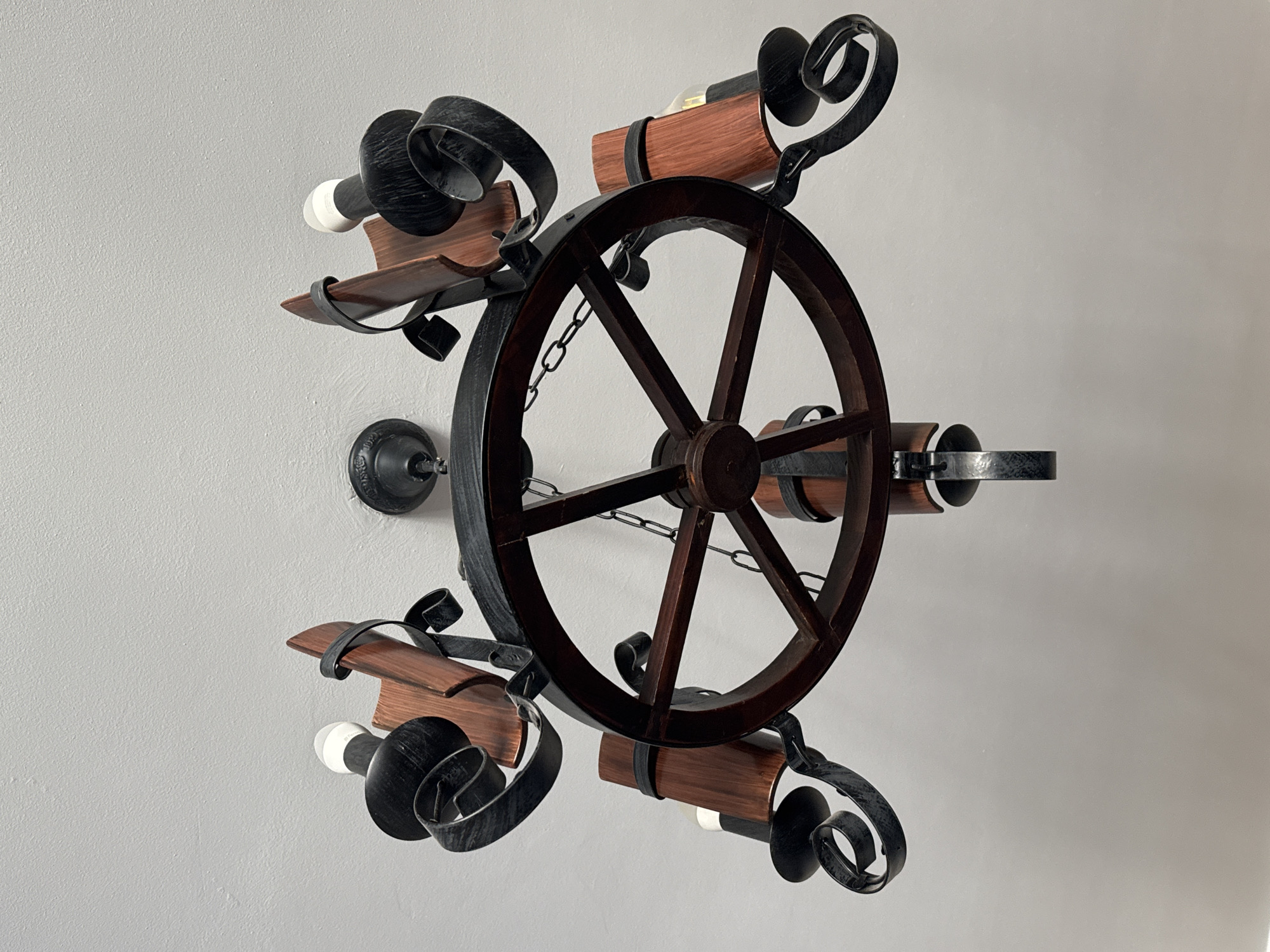 An overhead, low-angle, full shot captures a rustic, wagon wheel-style chandelier suspended from a white ceiling. The chandelier features a dark brown wooden wheel at its center, with metal spokes extending outwards. Attached to the spokes are five black metal candle holders, each with a wooden cylindrical shade. The candle holders are ornate with scrolling metalwork. The chandelier is lit by five LED bulbs. The scene is indoors, in a room with a white, textured ceiling. There are no people visible in the image. The location is unspecified, but the requested city of Bishkek, Kyrgyzstan, has been noted.