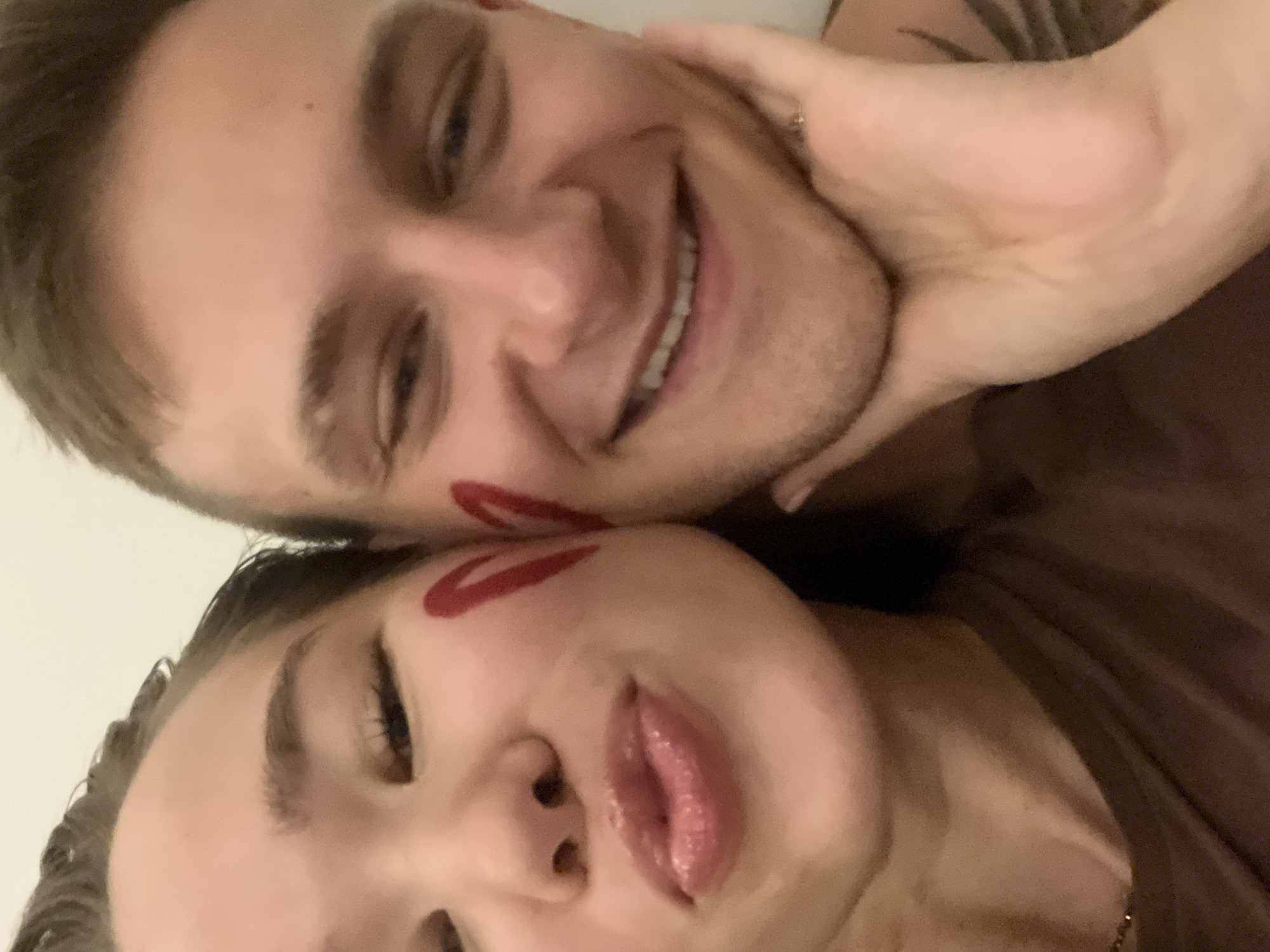 A close-up, tilted selfie features a smiling man and a woman. The man, positioned above, displays a broad smile with visible teeth. His arm is raised, and his hand gently cups the woman's cheek. The woman, located below, is also close to the camera. A red lipstick mark adorns her cheek. The background is blurred, suggesting a focus on the subjects.