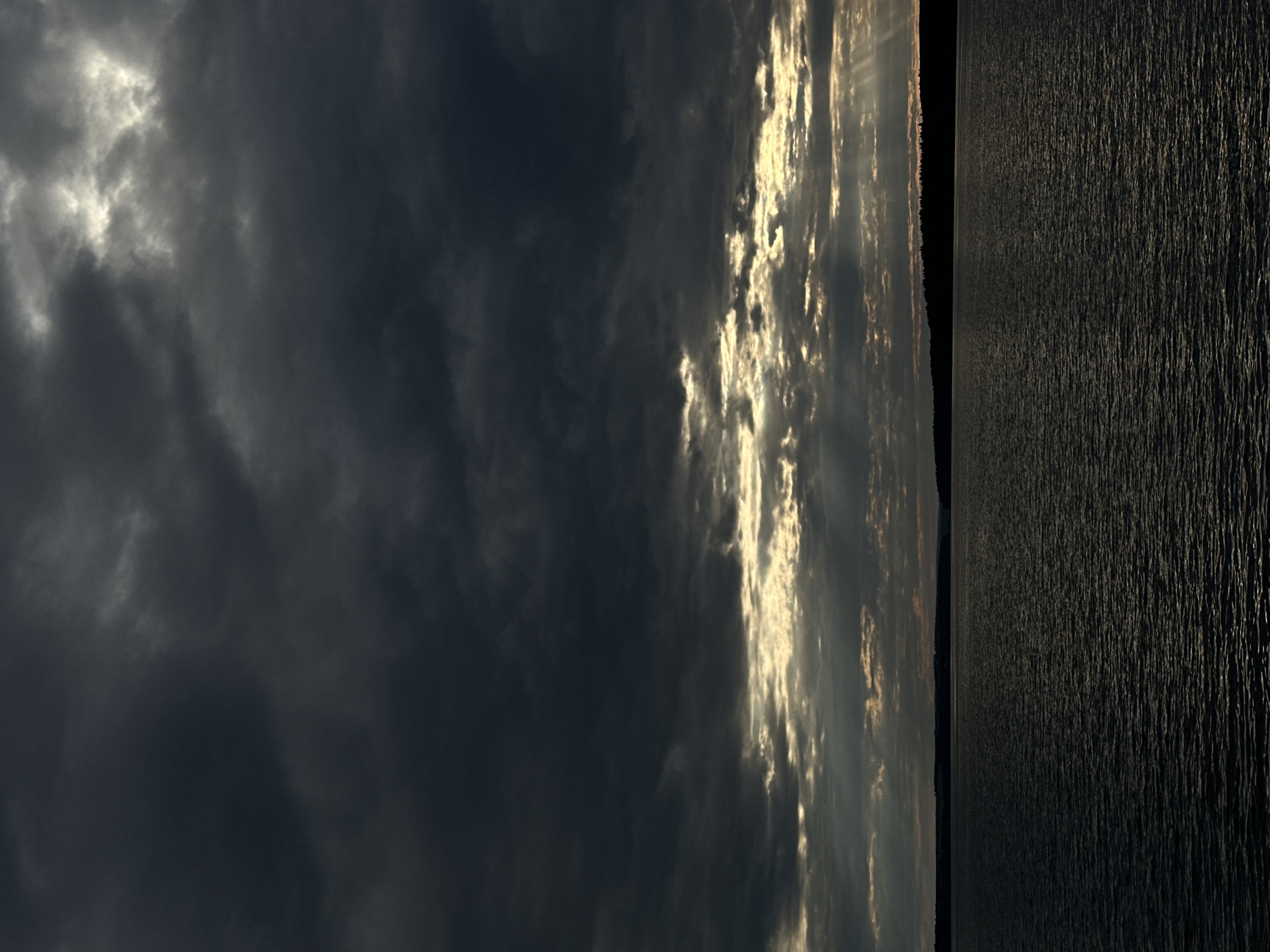 The image depicts a vast body of dark water under a dramatic sky filled with heavy, dark clouds. Sunlight pierces through breaks in the clouds, casting distinct rays onto the water. A silhouetted treeline forms the horizon in the distance. The scene is likely a natural landscape, possibly a lake or sea, with no visible human presence or man-made structures. The Votkinsk Reservoir, located in Votkinsk, Russia, is a large body of water that fits this description.