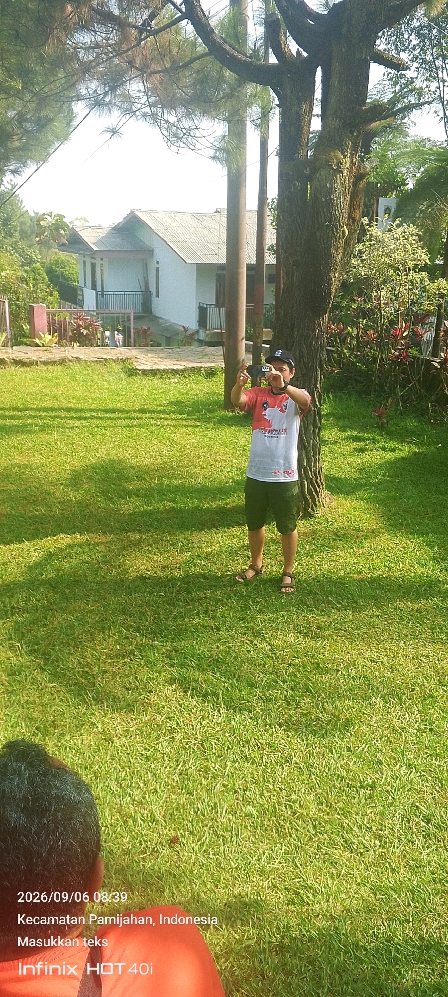 A male is standing in a grassy area next to a large tree, holding a mobile phone to take a picture. He is wearing a red and white t-shirt, green shorts, and brown sandals. In the background, there is a white house with a grey roof and a black railing in front of it. The scene is set in Pamijahan, Indonesia, on a sunny day, as indicated by the shadows on the grass. The lower portion of the image shows a close-up of another person's head and shoulders, wearing an orange shirt. A timestamp and location are visible in the bottom left corner: "2026/09/06 08:39, Kecamatan Pamijahan, Indonesia".