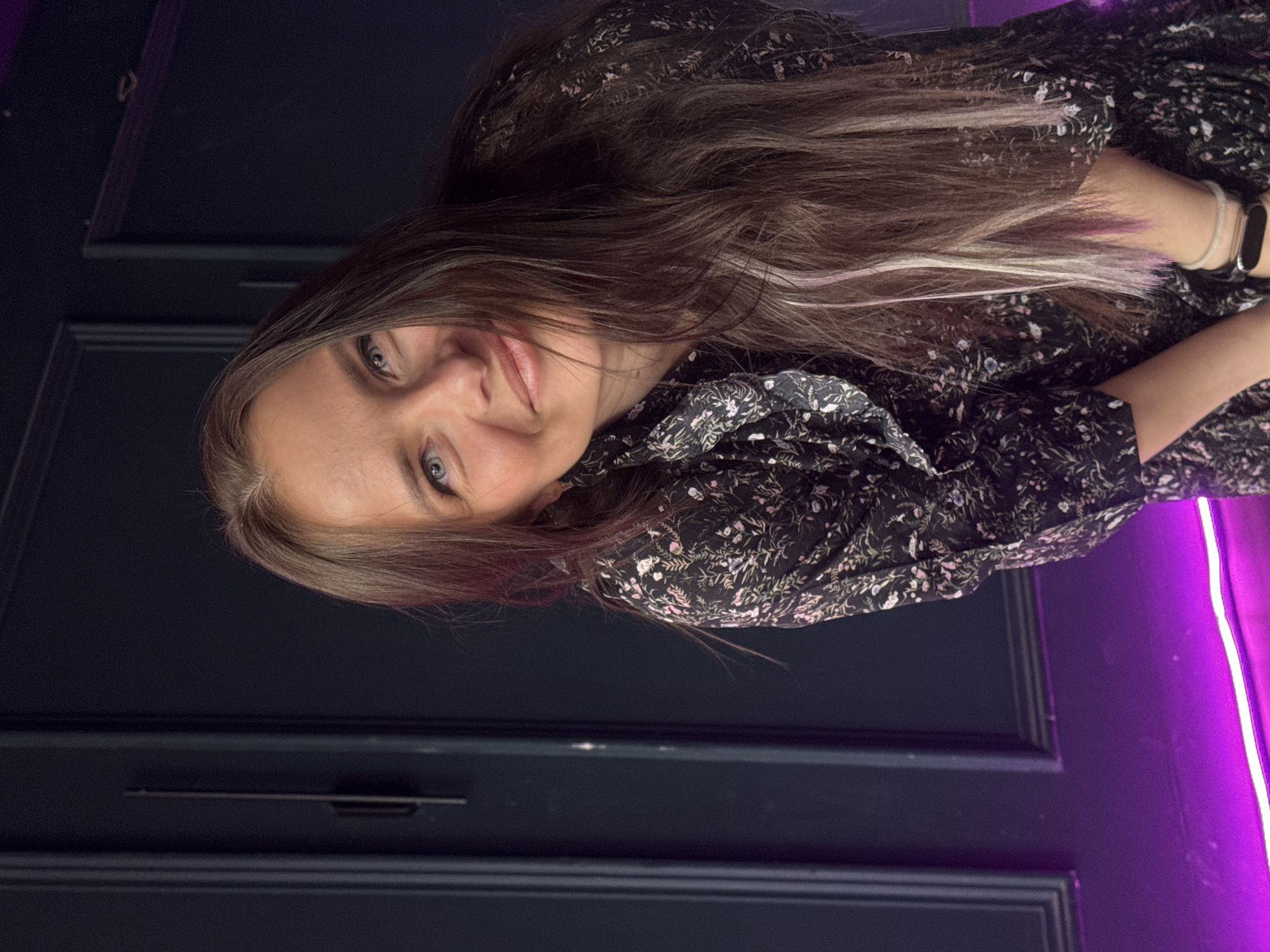 A woman with long brown hair is pictured indoors in a close-up shot. She is wearing a dark, floral-patterned dress. She is smiling slightly and facing the camera. Behind her is a dark blue paneled wall and a vertical strip of bright purple light. There is a pull handle visible on the wall. The location is Chaikovsky, Russia.