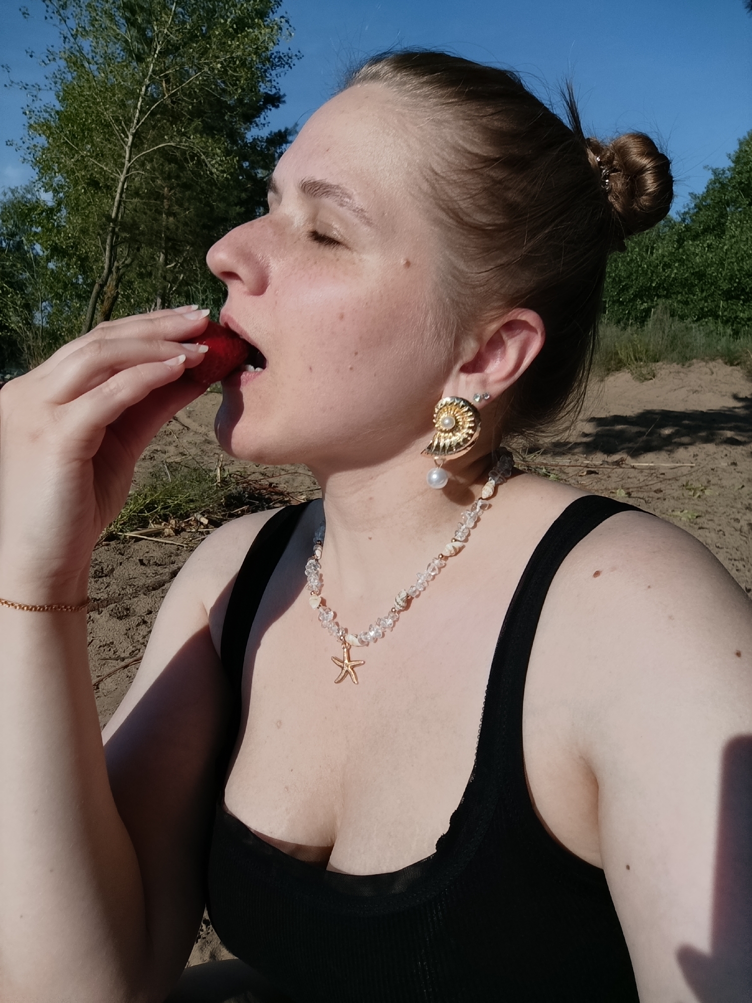 A close-up profile shot captures a woman with fair skin, freckles, and light brown hair pulled into a bun. She is holding a red strawberry to her mouth, eyes closed, appearing to enjoy the fruit. The woman is wearing a black tank top, a beaded necklace with a starfish pendant, and a large, shell-shaped earring with a pearl. A delicate gold bracelet is visible on her left wrist.
The background suggests an outdoor, natural setting. A bright blue sky is visible above, with green trees and foliage in the distance. The ground appears to be sandy or dirt, with some dry grass and twigs. The lighting indicates it is daytime, likely sunny, with shadows cast across the scene.
The overall mood seems relaxed and peaceful, perhaps a moment of enjoyment during an outing in nature. There is no visible text in the image. The location context provided is Подстёпки, Russia.