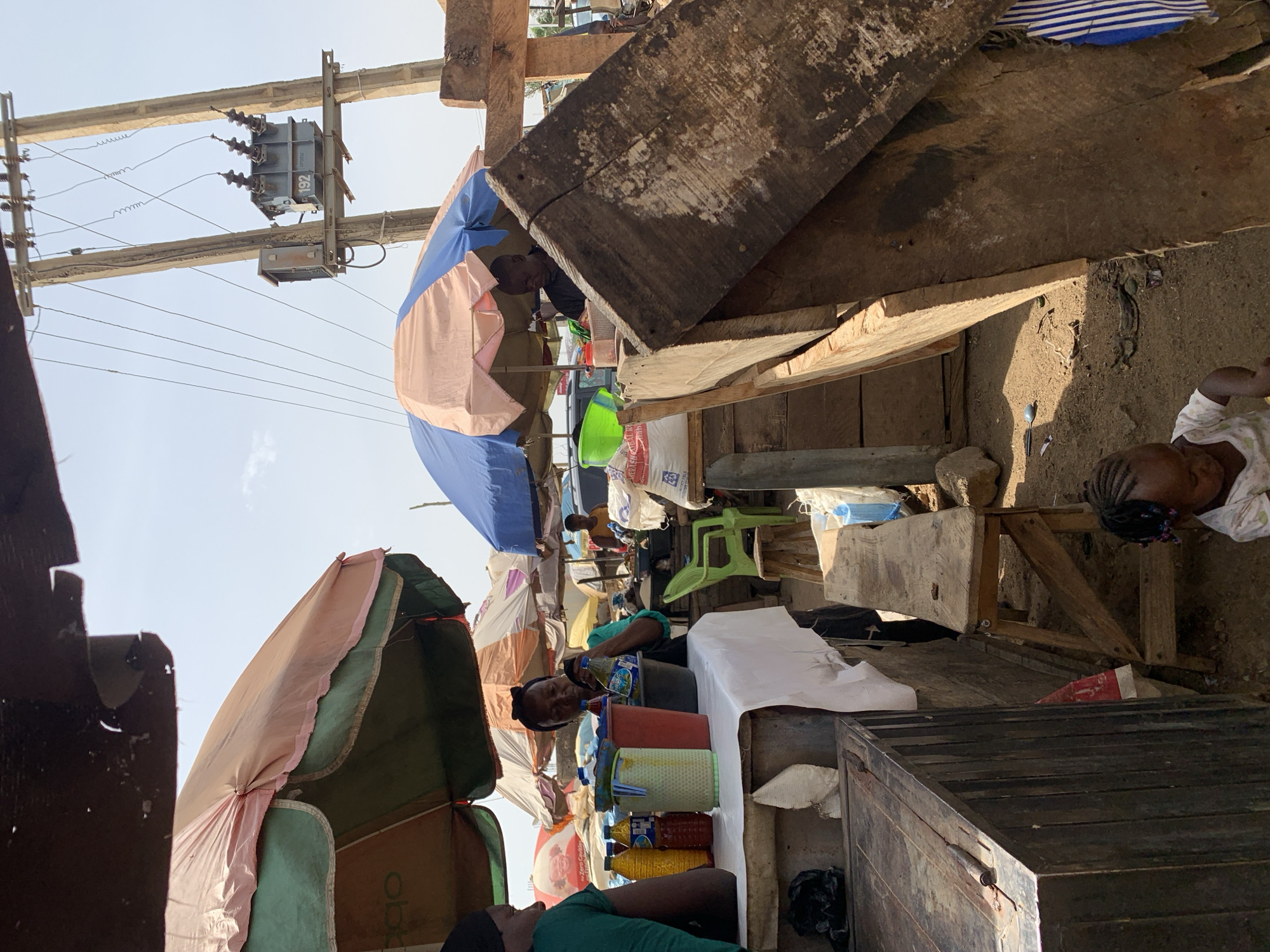 The image depicts a bustling street scene in Rafin Par, Nigeria. The setting is an outdoor market with stalls shaded by colorful umbrellas. Multiple people are present, including vendors and customers. A woman in a green shirt lies on the ground, while another woman tends to containers of liquid on a table. A child with braided hair is seen to the side. A wooden structure, utility pole, and transformer are visible above the stalls, with power lines crossing the sky. The overall environment is crowded and dusty.