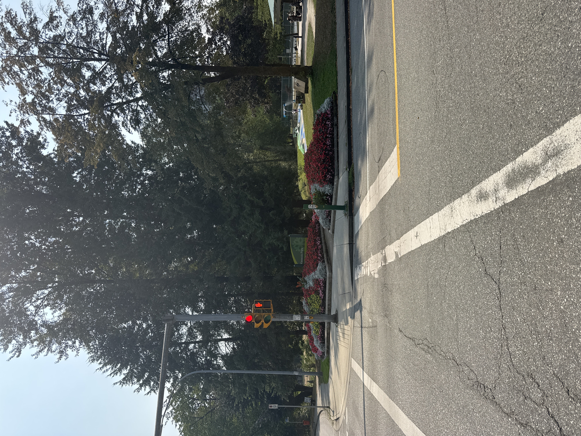 The image shows a street intersection with a pedestrian crossing and a traffic light. The traffic light is red, and a red hand symbol indicates that pedestrians should not cross. A sign for "Keswick Park" is visible in the background, along with trees, flower beds, and a playground. The street is paved with asphalt and has white and yellow lane markings. The weather appears to be sunny, with a clear blue sky. The location is identified as Burnaby, Canada. There are no people clearly visible in the image, only a few figures near picnic tables in the park. The scene is an ordinary urban streetscape with park amenities.