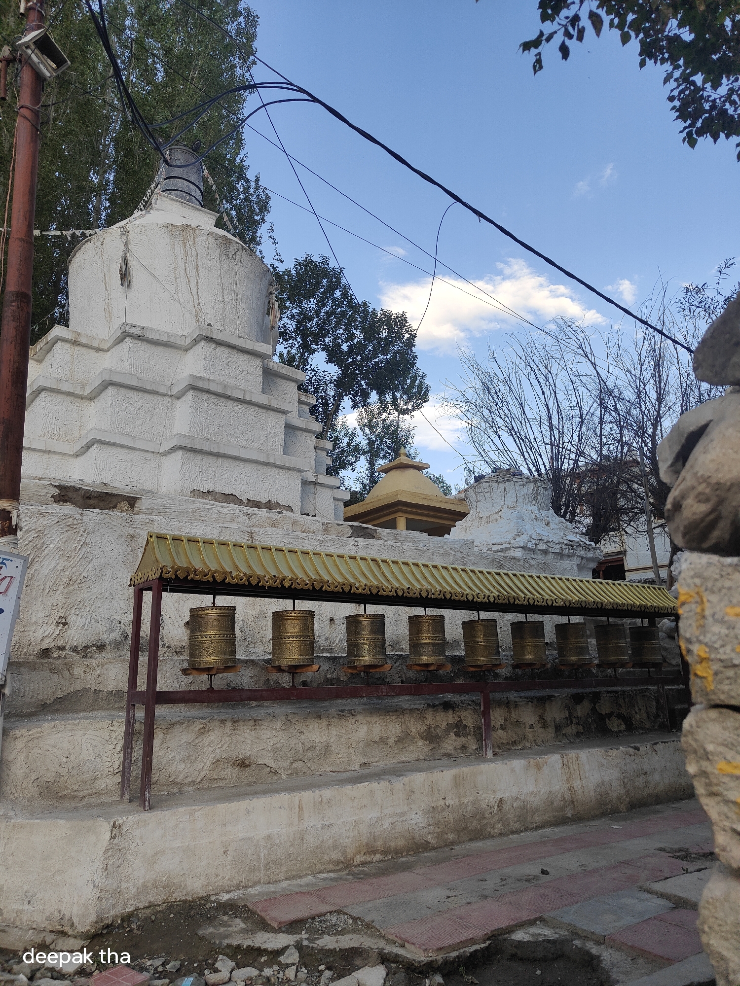 The image depicts a row of golden prayer wheels lined up beneath a decorative yellow awning. Behind the prayer wheels, a white stupa with tiered levels is visible, set against a backdrop of trees and a blue sky with scattered clouds. A brown utility pole with numerous electrical wires extends into the frame from the left. The ground in front of the prayer wheels is paved with reddish-brown tiles. The scene appears to be set in Leh, India, a region known for its Buddhist monasteries and cultural heritage, suggested by the presence of the stupa and prayer wheels.