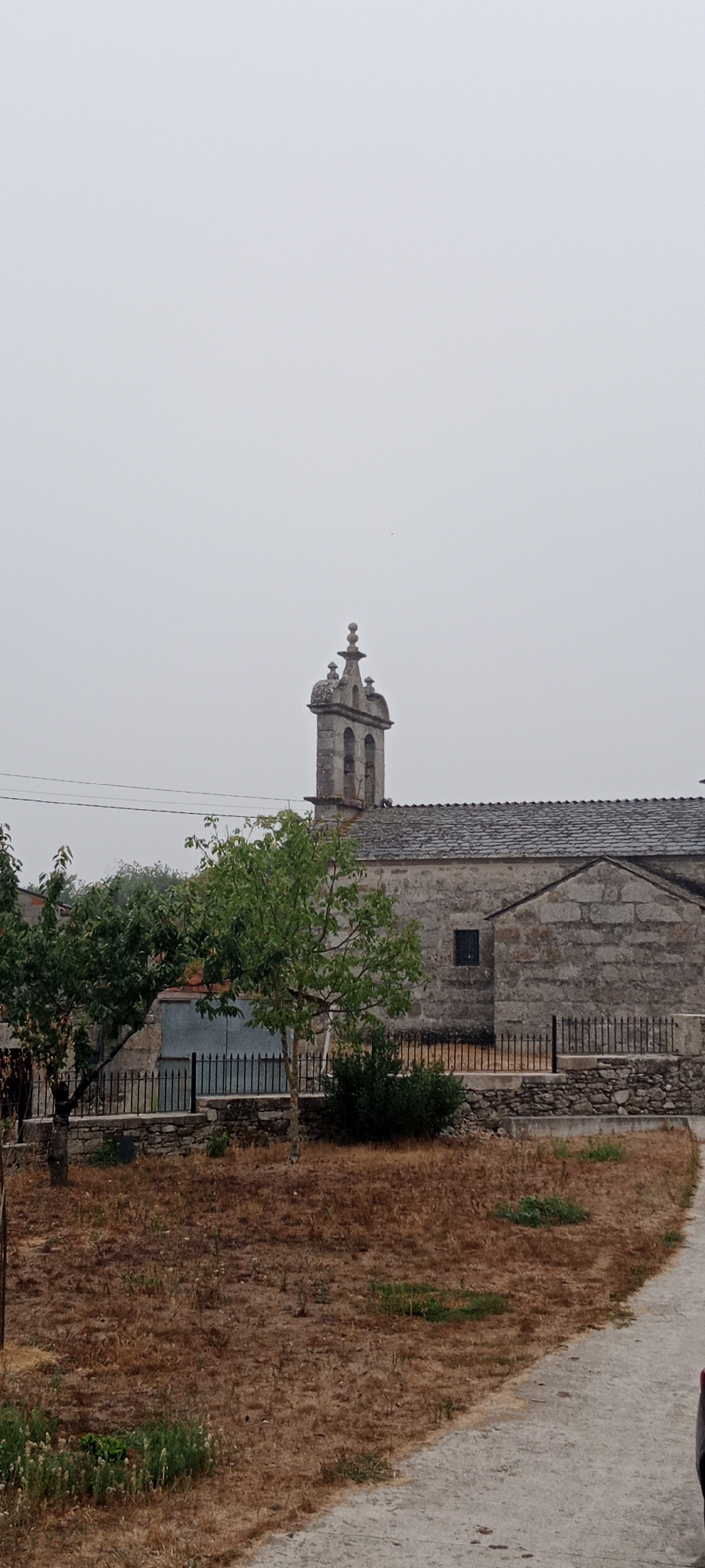 The image depicts a stone church with a bell tower in Adai, Spain. The church has a slate roof and a simple, rustic architectural style. In front of the church, there are two trees with green foliage, and a patch of dry, brown grass covers the foreground. A dirt path or road is visible on the right side of the image. The sky is overcast and grey, suggesting a cloudy day. No people or animals are visible in the scene.
