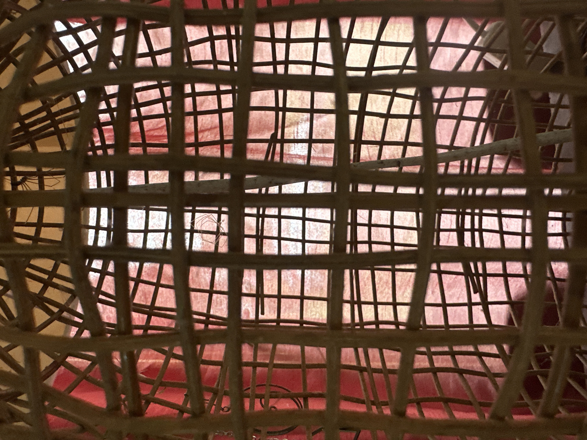 The image shows a close-up view through a woven bamboo cage. The cage's structure creates a grid-like pattern across the frame. Behind the cage, a red fabric is visible, with some areas appearing lighter or shadowed. The lighting suggests the photo was taken indoors, possibly with a light source behind the fabric. There are no people or obvious animals present. The location is not specified in the image.