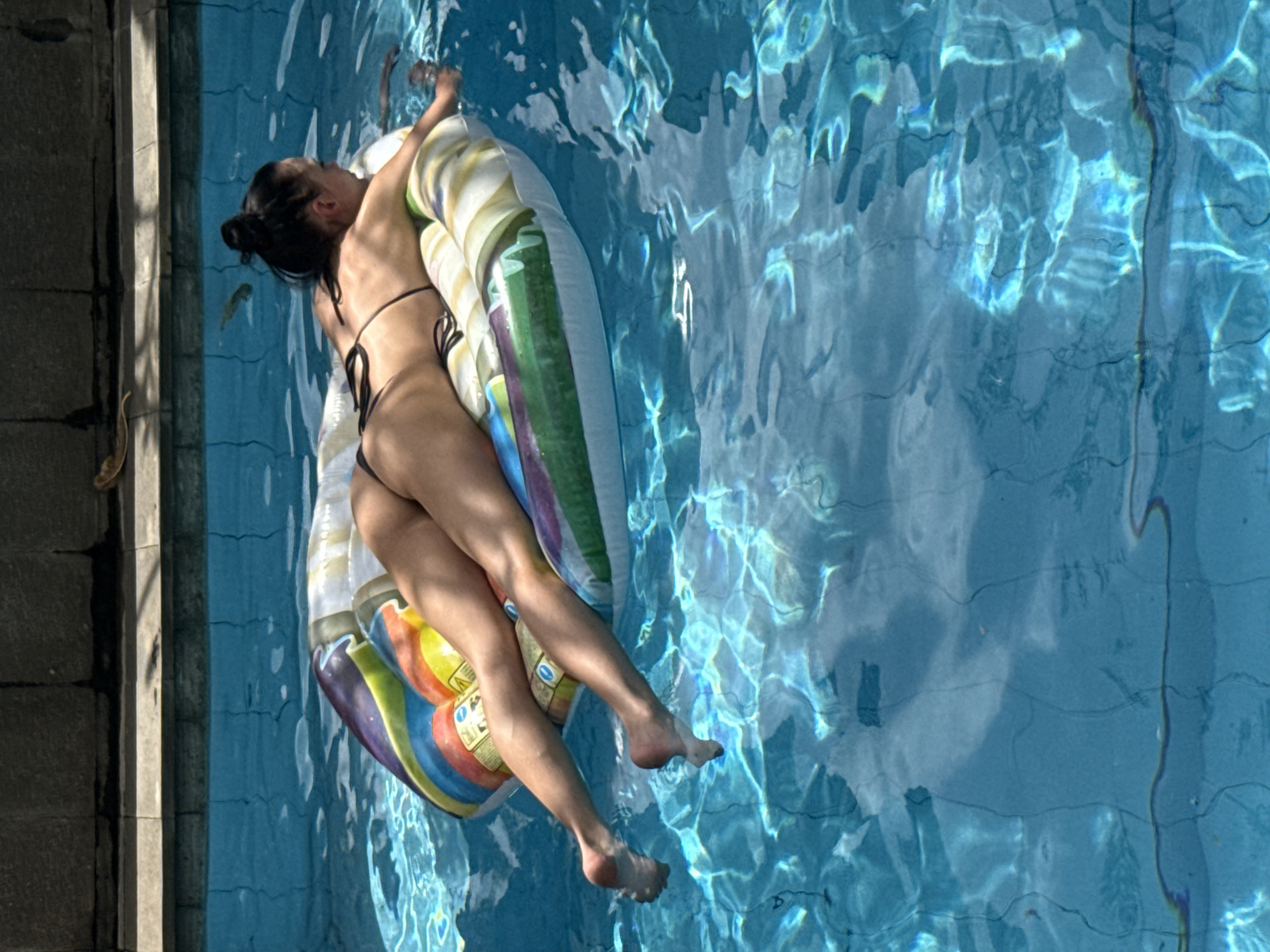 A woman in a black bikini lies on an inflatable raft in a swimming pool. Her body is slender, and her hair is pulled back in a bun. The raft is multicolored, with white, green, yellow, orange, red, purple, and blue stripes. The pool water is clear and blue, with reflections of light creating patterns on the surface. The pool is surrounded by grey stone. The location is North Kuta, Indonesia.
