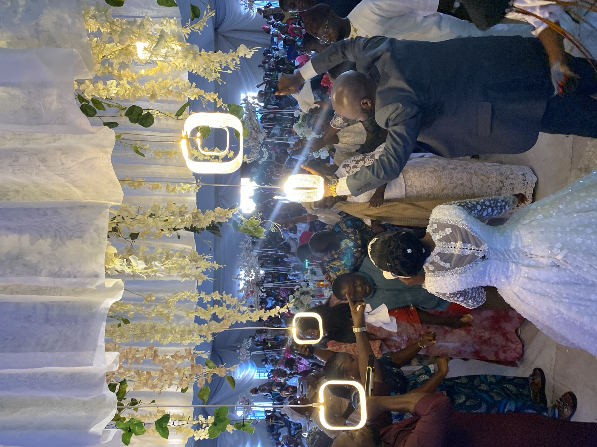 A rotated aerial view of a wedding in Vom, Nigeria. A bride in a white gown and a groom in a dark suit are surrounded by guests in colorful attire. The interior is decorated with white drapery, floral arrangements, and square-shaped pendant lights. Many guests are photographing the couple.