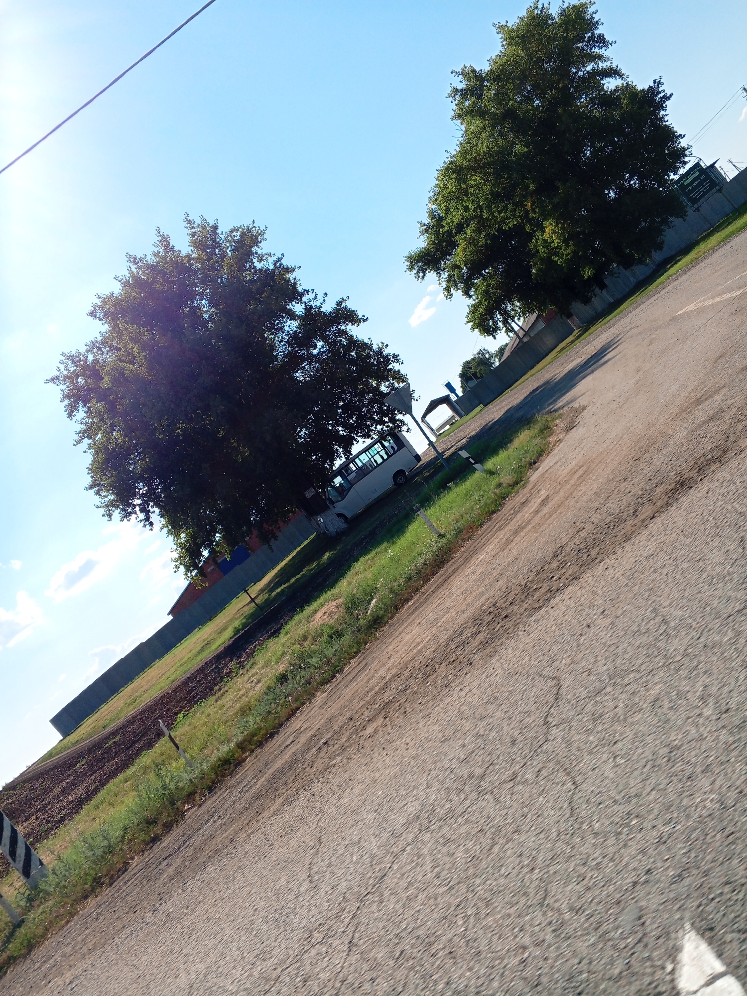 The image depicts an outdoor scene with a tilted perspective. A paved road, showing some cracks and dirt patches, occupies the foreground and extends diagonally across the frame. To the left of the road, there is a grassy area with some tilled soil. In the background, several trees with dense green foliage are visible. One of these trees, on the left side of the image, is partially backlit by the sun, creating a lens flare effect. Further in the background, there is a white bus parked on the side of the road, adjacent to a fence. Beyond the fence, buildings and structures can be seen. The sky is clear and blue with a few scattered clouds. A power line runs diagonally across the upper portion of the image. The overall impression is that of a rural or semi-urban environment on a clear day.