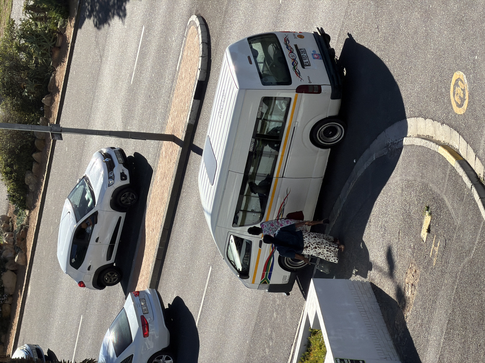 The image depicts an aerial view of a street scene, likely in Cape Town, South Africa, given the presence of a white Quantum minibus taxi (a common form of public transport in the region) and other cars with South African license plates. A white minibus taxi is parked near a concrete barrier with two individuals standing beside its side door. They appear to be interacting, potentially passengers boarding or disembarking. The ground is paved and shows signs of wear and repair. There is a white car parked parallel to the taxi, as well as another white car parked behind it. There is a circular painted traffic island on the road. Vegetation is visible along the side of the road. The lighting suggests a sunny day. The scene appears to be a relatively ordinary street setting in an urban area.
