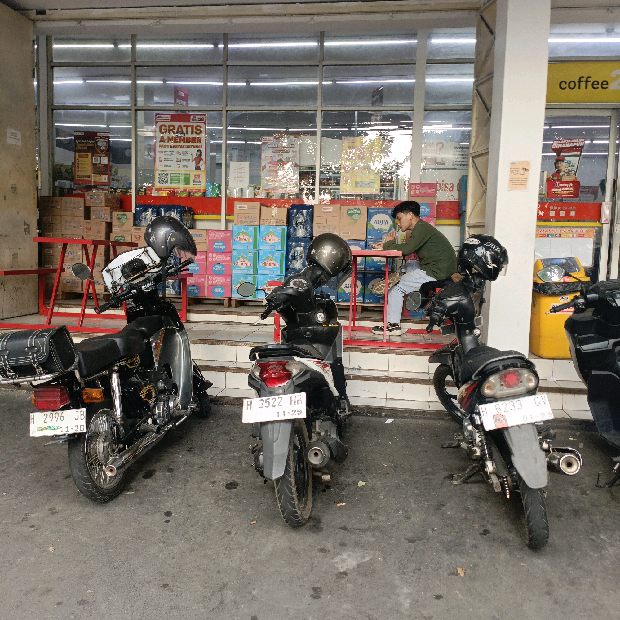 The image depicts a scene outside a storefront in Salatiga, Indonesia. In the foreground, three motorcycles are parked on a paved surface. The leftmost motorcycle is a classic model, black with chrome accents, a black saddlebag, and a helmet resting on its handlebars. The middle motorcycle is a smaller, modern scooter, gray and black, also with a helmet on its handlebars. The rightmost motorcycle is a black scooter with a helmet on its handlebars.
A young man is seated at a red table to the right of the middle motorcycle, looking down at his phone and holding a beverage. He is wearing a dark green long-sleeved shirt and light blue jeans. Behind the man and motorcycles, the storefront features large glass windows revealing shelves stocked with bottled water and other products. A prominent yellow sign with the word "coffee" is visible above the entrance on the right side of the image. The storefront is brightly lit with fluorescent lights. Boxes of goods are stacked on shelves and on the floor behind the motorcycles. The overall environment appears to be a commercial area, likely a minimarket or convenience store.