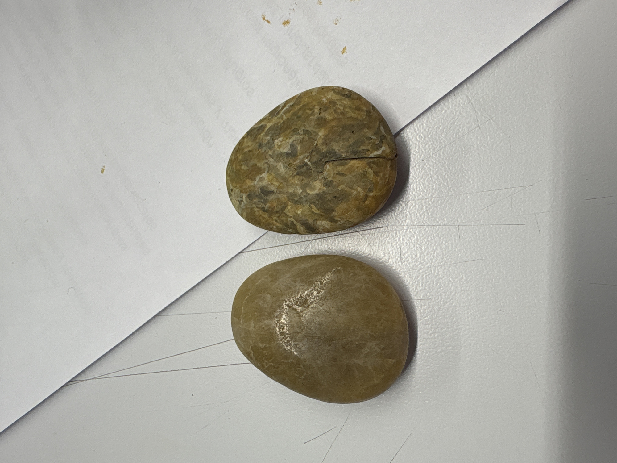The image shows two rocks placed on a white surface, part of which is covered by a sheet of white paper. The top rock is textured and has a crack running across it. The lower rock is smoother and more uniformly colored. Fine threads or lines are visible on the white surface. No information suggests the image was taken in Mexico City, Mexico.