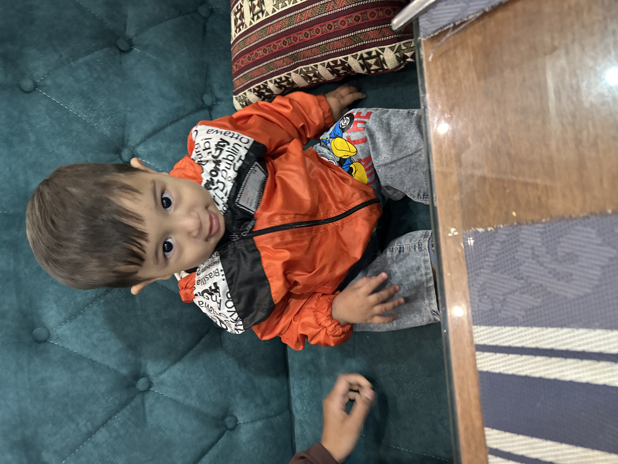 A child wearing an orange and black jacket and gray jeans sits on a teal, tufted couch. A decorative pillow rests behind the child. A hand of an adult is visible in the foreground. The location is inferred to be in or near Ақсу, Kazakhstan, based on the request.