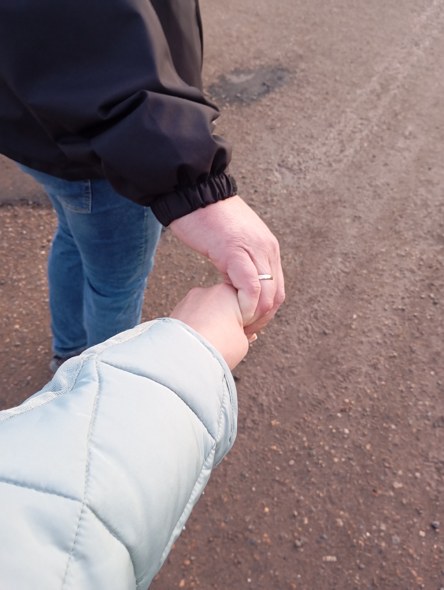The image presents a close-up, downward-angled view of two individuals holding hands while walking outdoors. In the foreground, the arm and hand of one person are visible, clad in a light blue, quilted jacket with distinct stitched patterns. This person's smaller, fair-skinned hand is clasped around the fingers of another individual's hand.
The second hand, appearing larger with a more mature texture, wears a simple gold-colored band on the ring finger. This hand belongs to a person partially visible in the mid-ground, who is wearing a black jacket with elasticated cuffs and blue denim jeans. Their lower body and feet are slightly out of focus, indicating movement.
The scene unfolds on an unpaved surface, likely a dirt path or gravel road, characterized by reddish-brown earth mixed with various small stones and pebbles. Patches of darker, possibly damp, ground are also visible. The lighting suggests it is daytime, with a soft, warm quality illuminating the ground. The central activity is the intimate gesture of holding hands, implying companionship, affection, or guidance between the two people. No text is visible within the image itself. The provided location context is Фоки, Russia.
