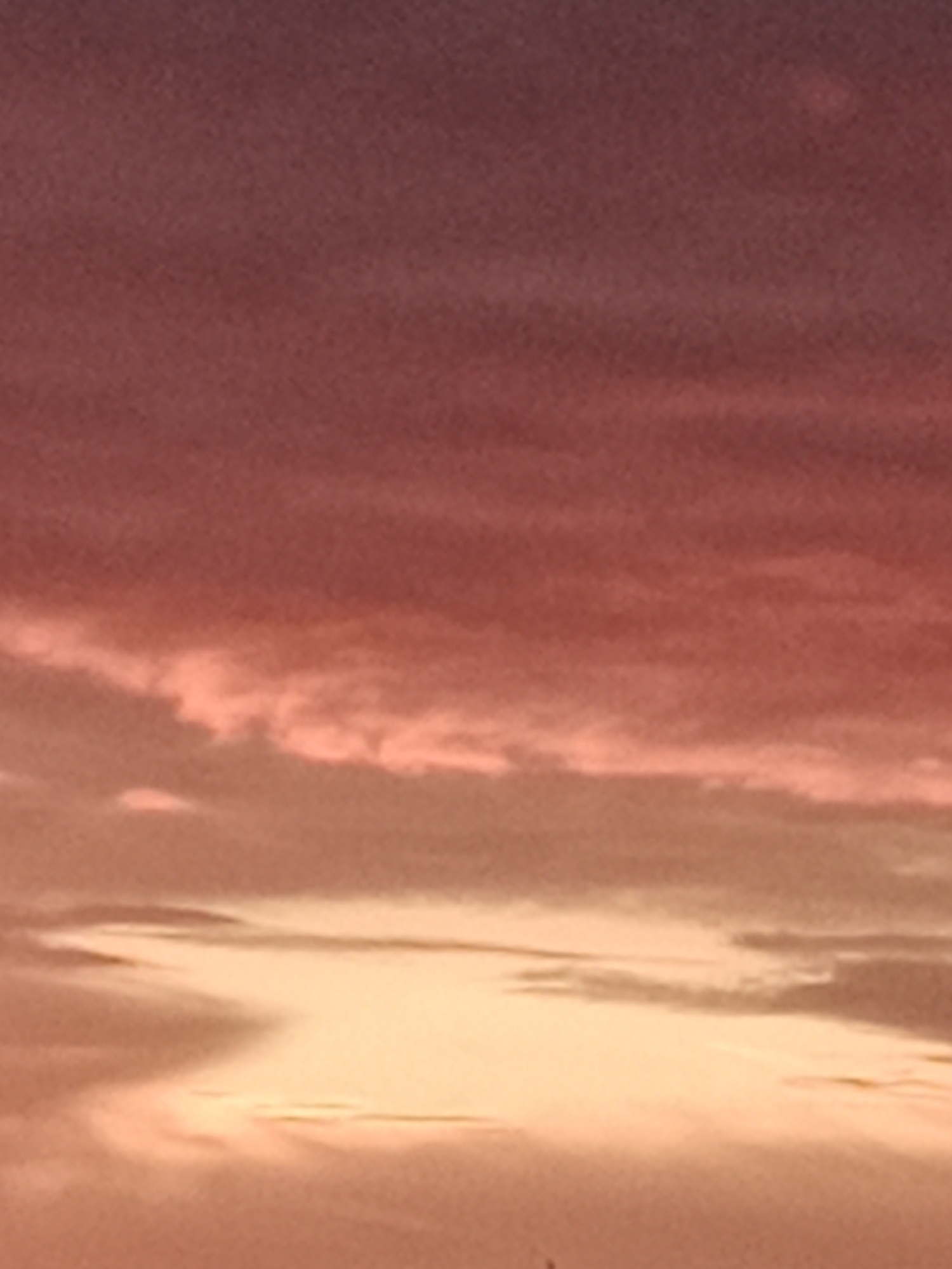 The image shows a sunset sky with varying shades of pink and orange. There are some clouds visible, with hints of sunlight peeking through. The lower part of the image is darker, suggesting the presence of a landscape or cityscape below the horizon, though no distinct features are visible. The scene is likely outdoors. The image does not contain any identifiable landmarks or specific details that would allow for pinpointing a location within Bydgoszcz, Poland.