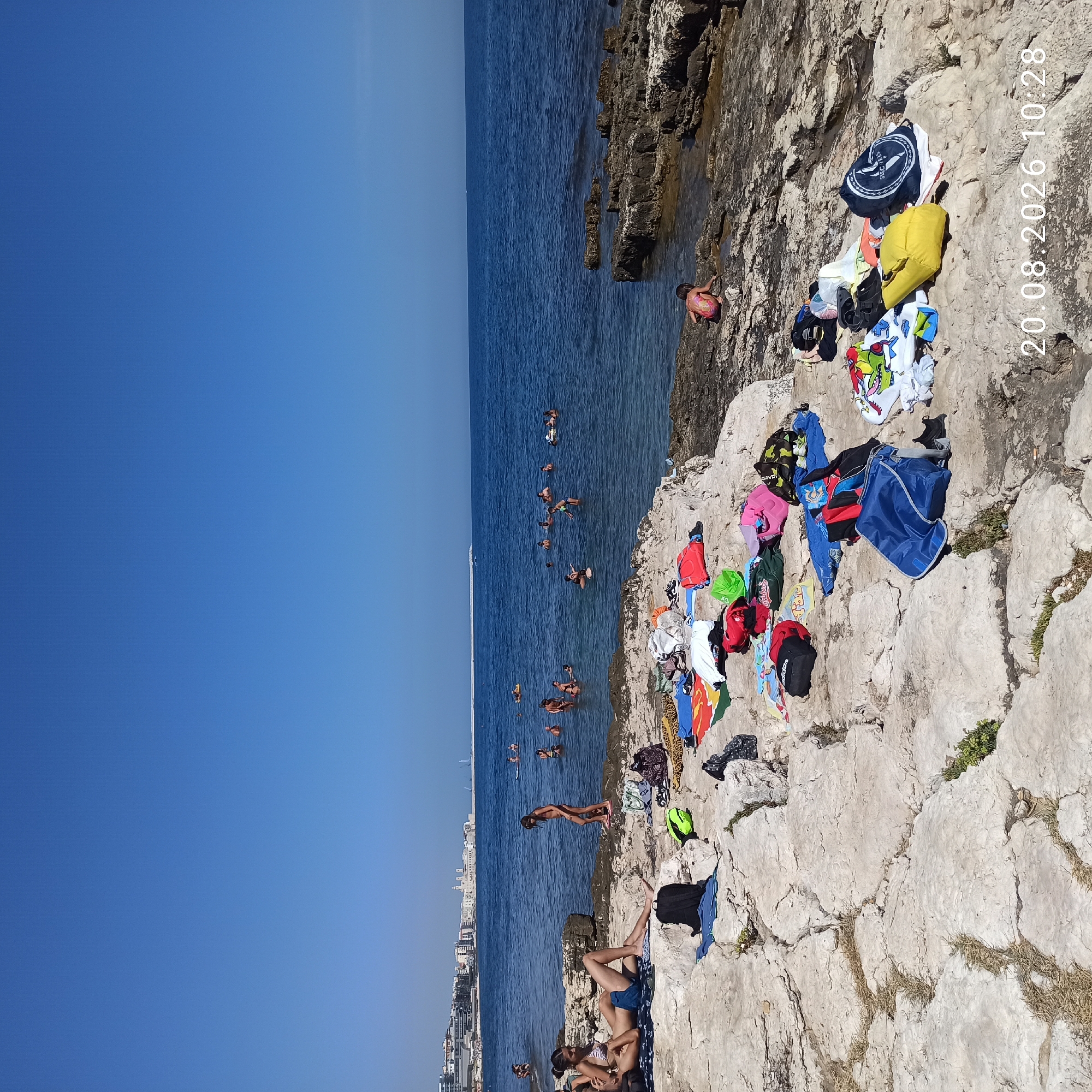 The image depicts a rocky coastline with a body of water in the background, suggesting a beach or seaside location. The water appears calm and blue, with several people swimming or wading in it. On the rocky shore, there are various items of clothing, towels, and bags spread out, indicating that people are sunbathing or relaxing. A few individuals are visible on the shore, including a young girl playing near the water's edge and a couple sitting on the rocks. In the distance, a city skyline with buildings and a lighthouse is visible, suggesting an urban setting adjacent to the coast. The overall scene conveys a leisurely summer day at the beach. The timestamp in the lower right corner indicates the date "20.08.2026 10:28".