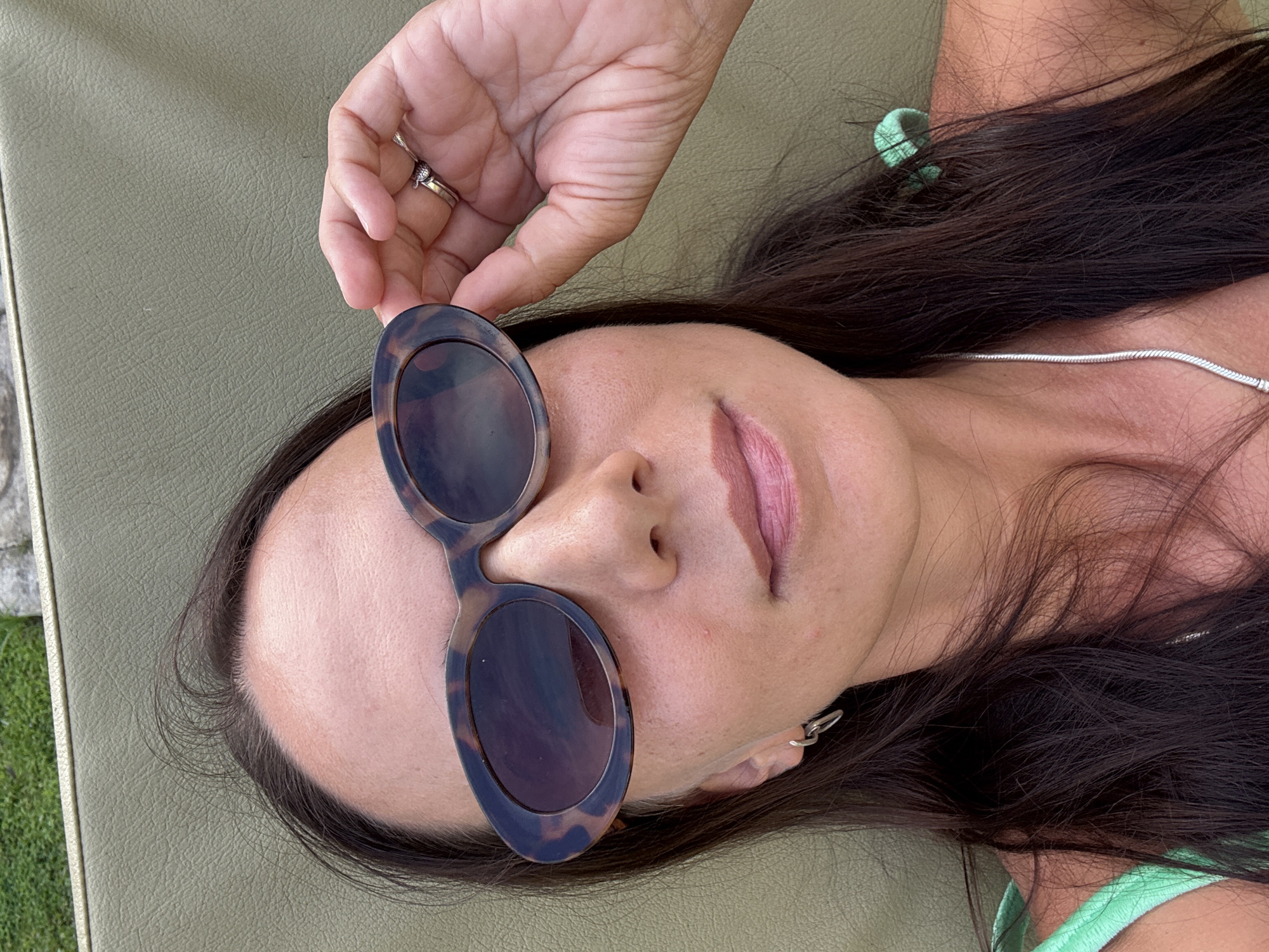 The image shows a woman lying on a green surface, possibly a lounge chair. She is holding a pair of sunglasses with a tortoiseshell frame partially in front of her face with her right hand, on which she wears a ring. She has dark hair, fair skin, and is wearing a silver necklace. The location is suggested to be Kuta Selatan, Indonesia, but there are no visual indicators to confirm this.