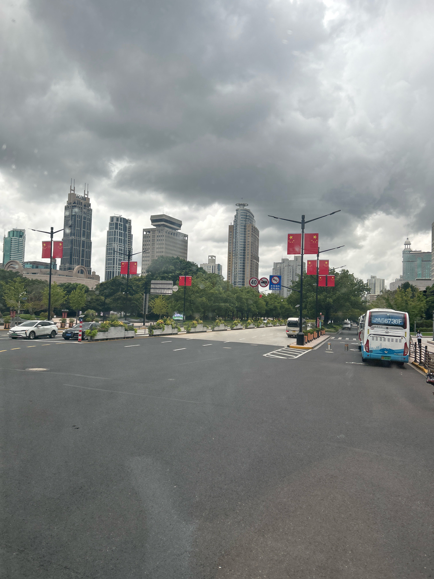 This is a medium shot of a street scene in Shanghai, China, under a cloudy sky. In the foreground, a paved road with white lane markings stretches towards a cityscape dominated by modern skyscrapers. Several Chinese flags, red with yellow stars, are prominently displayed on poles along the roadside, suggesting a patriotic or official setting.
Several vehicles are visible on the road. A white SUV is on the left lane, followed by a dark sedan. Further ahead, a light-colored van is seen. On the right side of the road, a blue and white bus with a license plate reading "A56736F" is partially visible. A person in dark clothing is standing near the bus.
The midground features a strip of green trees and manicured gardens separating the road from the buildings. Some streetlights are positioned along the road. The background is filled with a variety of tall buildings, including a distinctive skyscraper with a unique spire at its peak. Other buildings exhibit modern architectural designs, some with visible windows and facades. A traffic sign with "40" and other symbols is also present. The overall impression is an urban environment, possibly a significant boulevard or plaza, with a sense of order and civic pride conveyed by the flags.