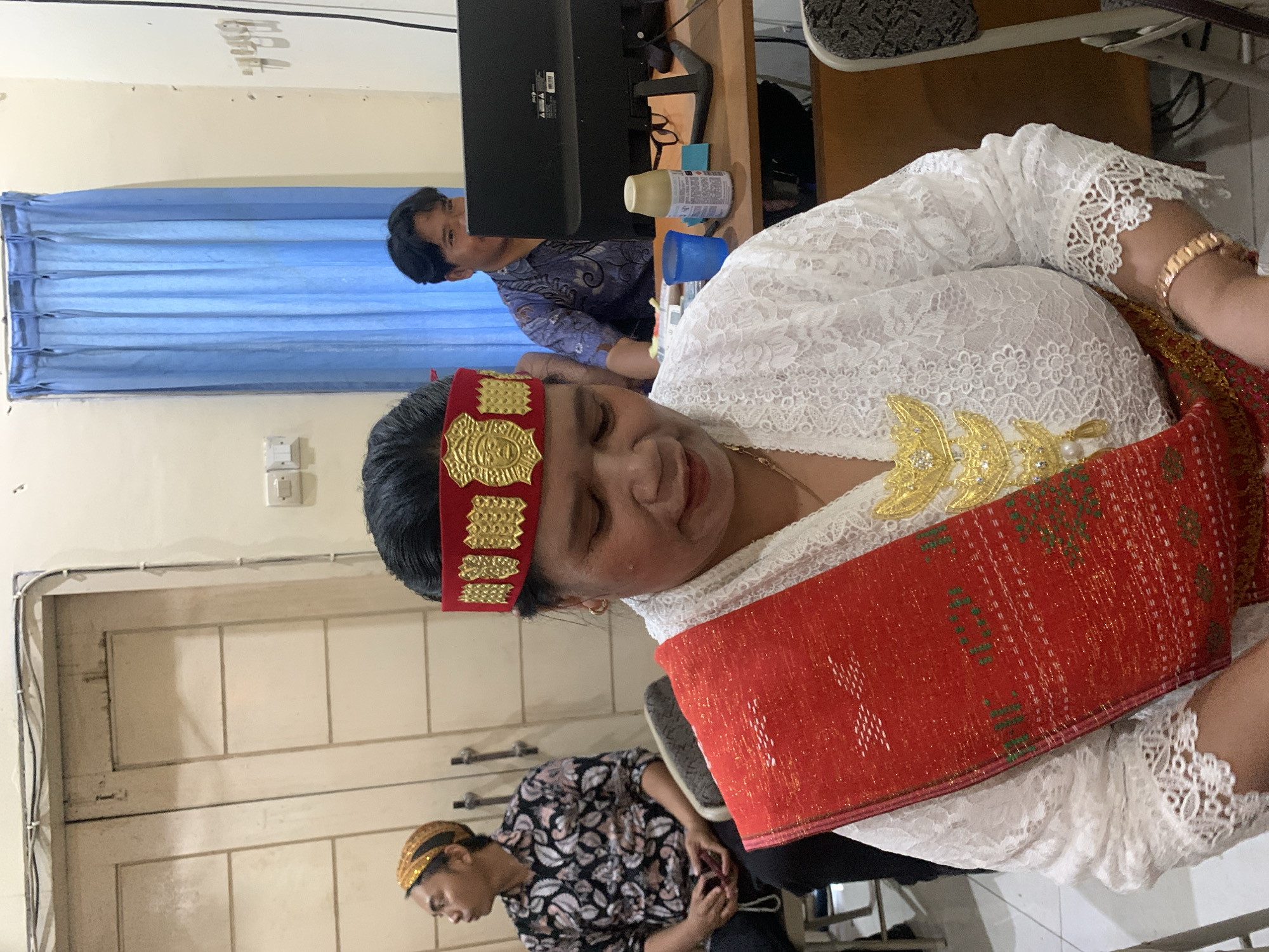 In Dukuh Mojo, Indonesia, a woman in traditional attire, featuring a red and gold headdress and a white lace top with a red sash, sits in the foreground. She wears gold jewelry, including a necklace and bracelet. To her left, a man in a patterned shirt and traditional headwear sits in a chair. In the background, another man, wearing a patterned blue shirt, is seated at a desk with a computer monitor. Blue curtains hang on the wall behind him. The scene appears to be indoors, possibly an office or meeting room, with closed doors visible on the left.