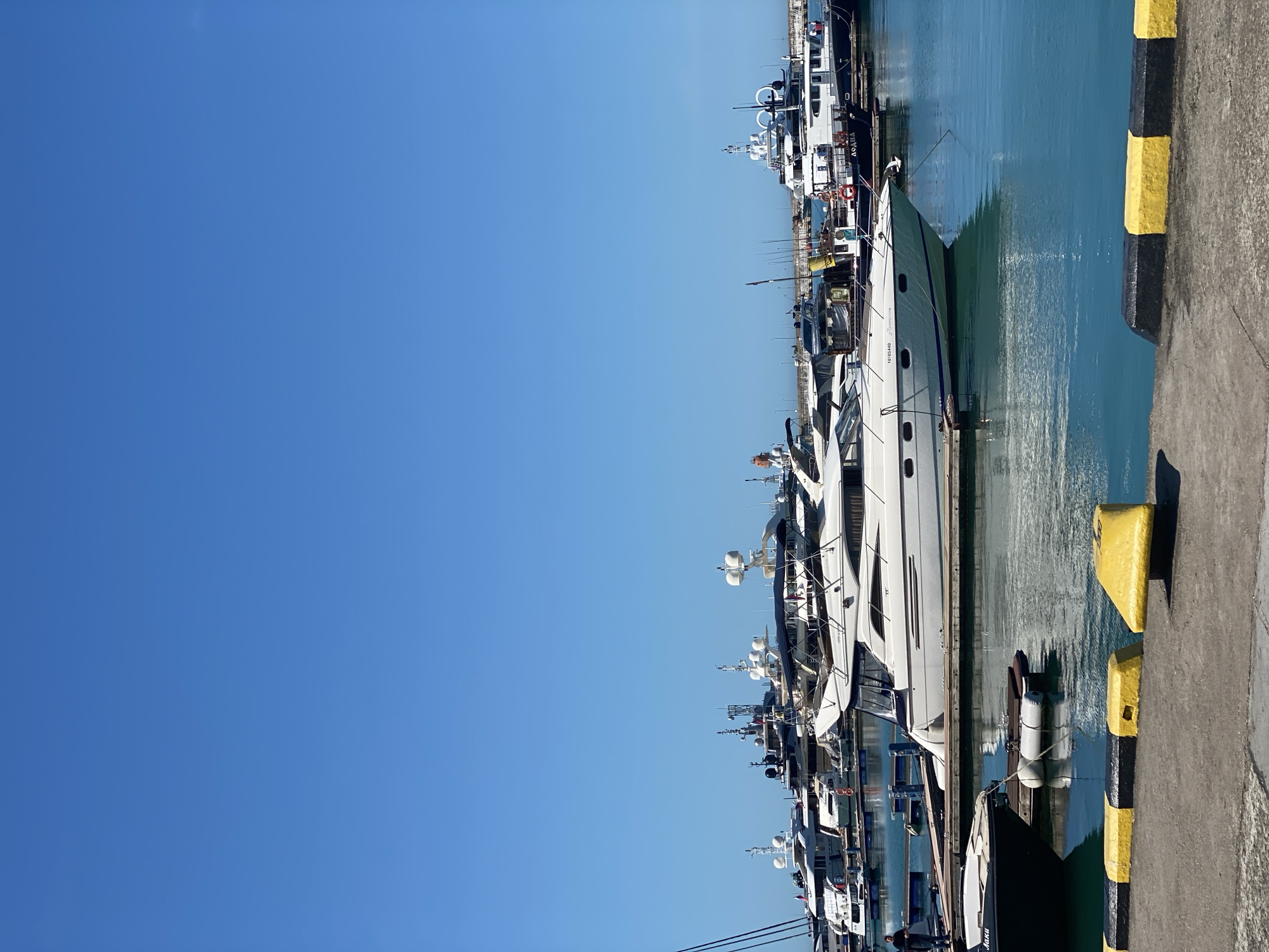 The image displays a bustling marina in Sochi, Russia, under a clear, bright blue sky. Numerous luxury yachts and motorboats of varying sizes are tightly moored side-by-side along concrete docks. The water in the harbor is calm and a serene blue-green, reflecting the hulls of the vessels and the sky above.
The boats are predominantly white or light-colored, featuring multiple levels, windows, antennas, and radar domes, indicative of modern recreational and cruising vessels. In the foreground, a prominent white yacht shows the identification number "1015640" and the name "Transamerica" clearly visible on its hull. Further back, another boat bears the name "ΛΟΔΗΔΙΑ" (LODHIA), and a different vessel displays "ЯНКИ" (YANKI) on its side.
To the right of the frame, a concrete pier edge is visible, adorned with distinctive black and yellow striped safety bollards. White mooring buoys or fenders are scattered in the water, secured near the docked boats. The scene is bathed in bright daylight, suggesting a sunny mid-day with no clouds. No prominent people are visible, emphasizing the boats and the peaceful marina environment. The overall impression is one of tranquility and maritime leisure.