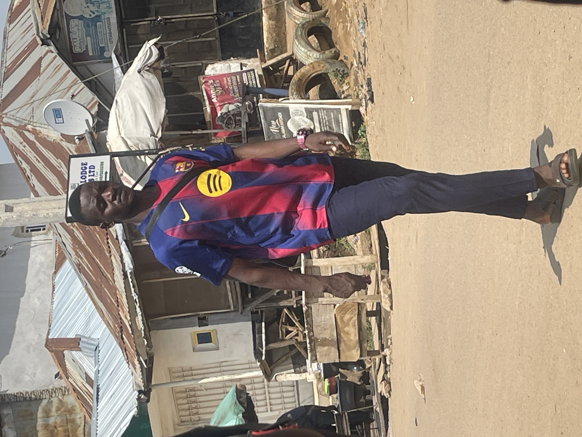 A full-body, eye-level, outdoor shot shows a young Black man walking down a dusty, paved street in Abuja, Nigeria. He is wearing a blue and red striped FC Barcelona soccer jersey with a yellow Spotify logo on the chest, dark blue pants, and dark sandals. A black strap bag is slung across his chest. His left hand is clenched, and he is holding a crumpled purple cloth. In the background, there are several buildings with corrugated metal roofs, some appearing weathered and rusty. Signs for "PAULA'S," "Annie's Bites," and "New Calabar Kitchen" are visible. A satellite dish is mounted on one of the buildings. Several old tires are stacked on the side of the road. The ground is covered in sand and debris. The lighting suggests it is a bright, sunny day.