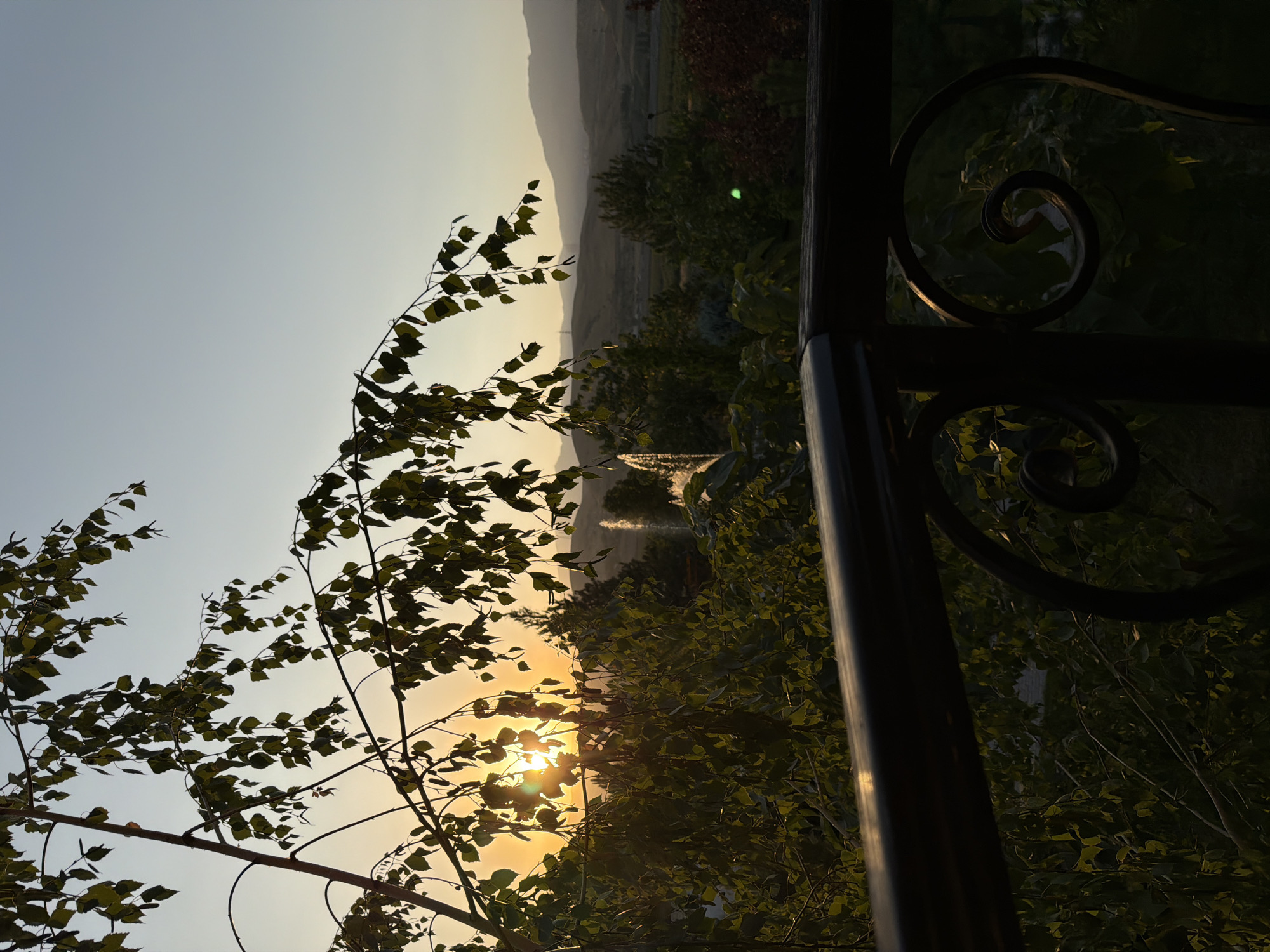 A serene sunset scene unfolds over a landscape in Karankultugay, Uzbekistan, viewed from a balcony. The foreground is dominated by the dark silhouette of a balcony railing with decorative scrollwork. Beyond the railing, lush green trees fill the midground, their leaves catching the warm, golden light of the setting sun. The sun itself is partially obscured by the branches and leaves of a tree in the upper left portion of the frame, casting a soft glow across the scene. In the distance, rolling hills fade into a hazy horizon under a pale blue sky. A fountain can be seen in the middle ground, its water catching the light. The overall atmosphere is peaceful and tranquil.