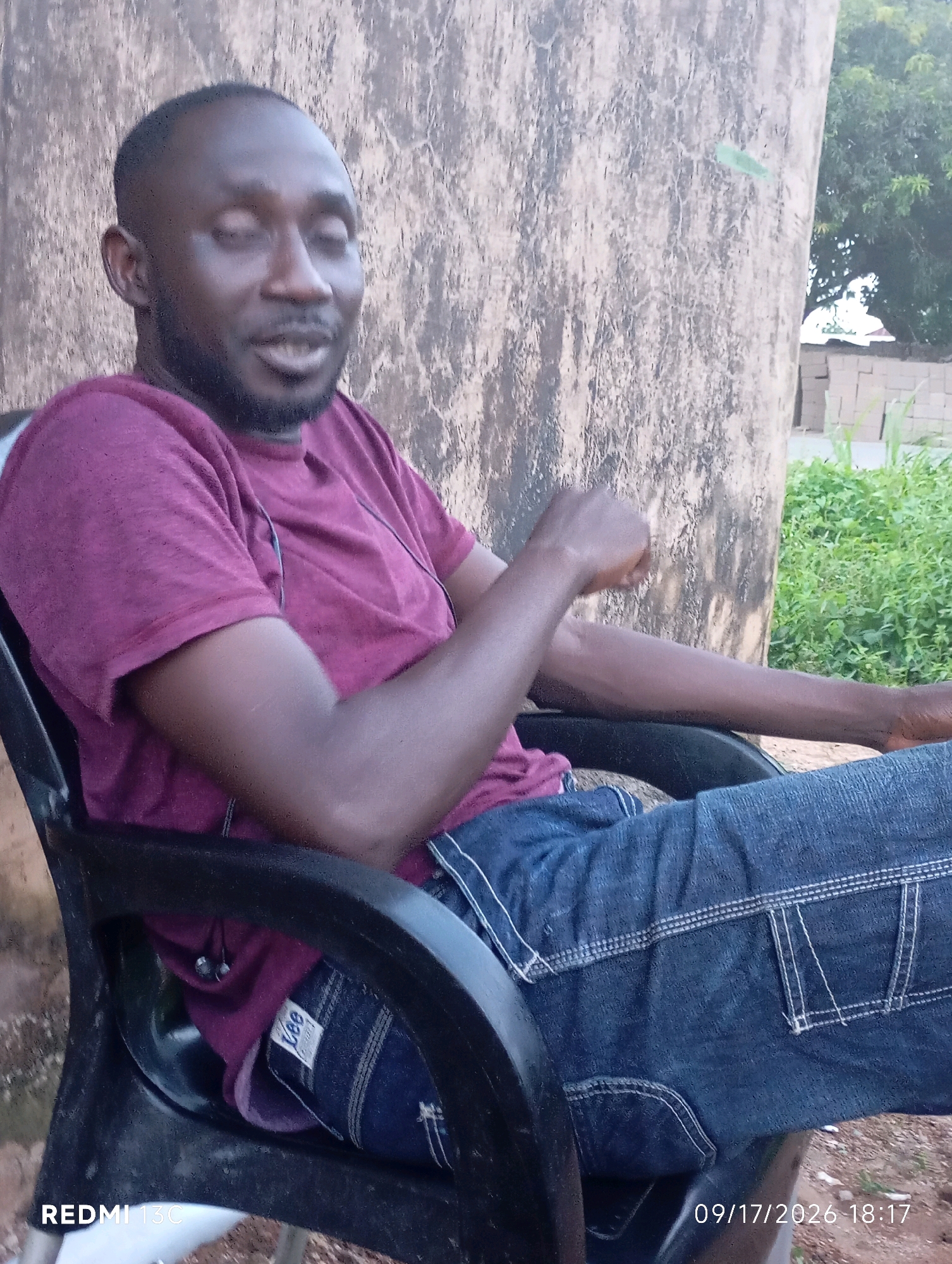 A medium shot photograph shows a Black man sitting in a black plastic chair outdoors, with his body angled towards the right. He has short dark hair, a short beard, and is wearing a maroon t-shirt and dark blue jeans with white stitching. He is looking to his left with his eyes closed and mouth slightly open. His right arm is bent with his hand in a fist near his chest, and his left arm is resting on his lap. A pair of dark earbuds are visible in his ears, with wires running to his shirt. Behind him, a large textured wall, possibly a building or a tree trunk, dominates the background. To the right of the wall, there is green foliage and a glimpse of what appears to be a brick building under construction. The bottom right corner of the image shows a date and time stamp: "09/17/2026 18:17". The text "REDMI 13C" is visible in the bottom left corner. The scene appears to be in a rural or semi-urban setting in Shendam, Nigeria.