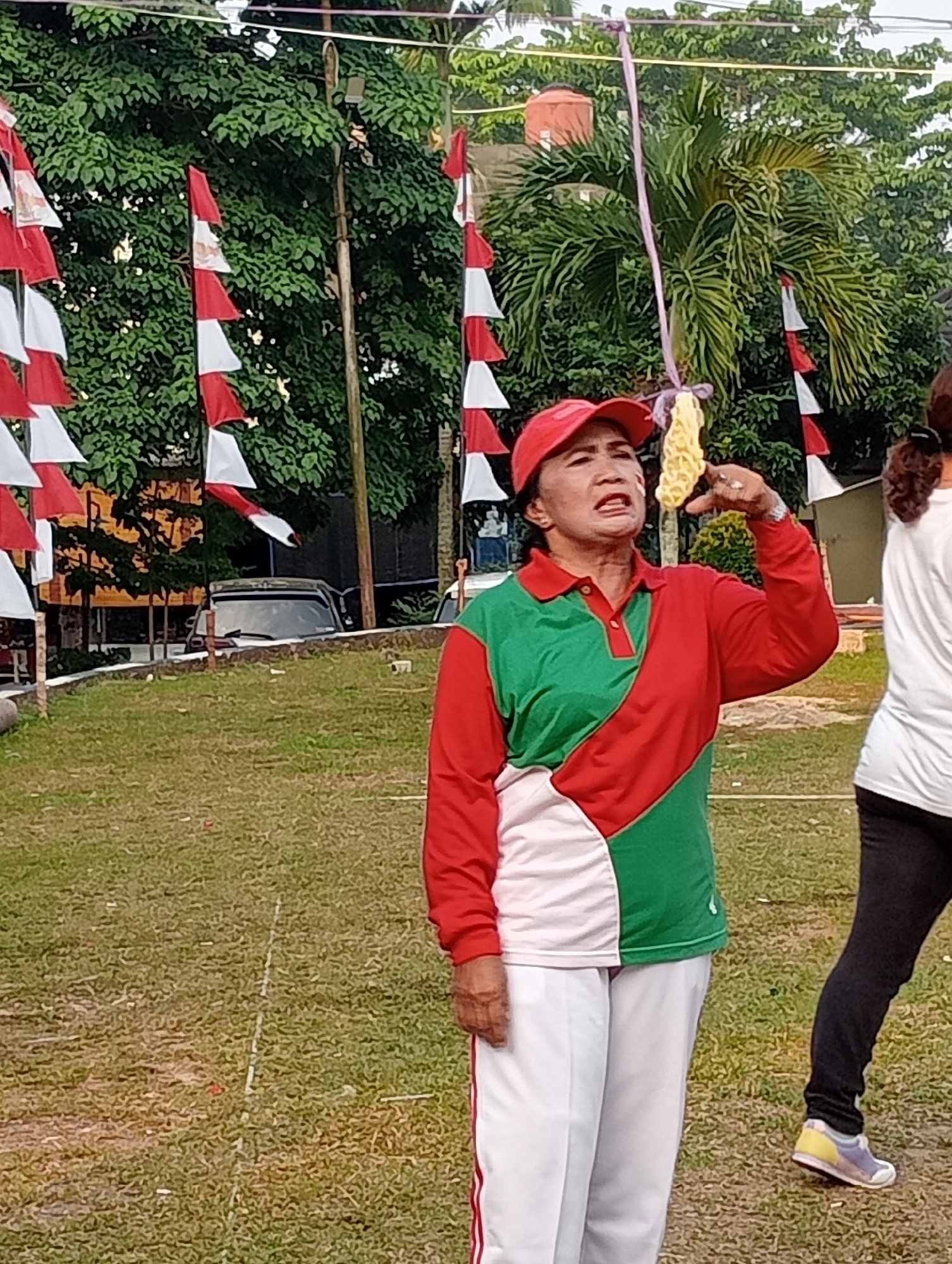 A woman in a red cap and a red and green shirt is playing a game outdoors. She is holding a yellow cracker on a string and trying to eat it with her mouth. She is wearing white pants with red stripes. There is another person partially visible on the right side of the image. The game is taking place on a grassy field with trees and Indonesian flags in the background. There are also some buildings and a car visible in the distance.