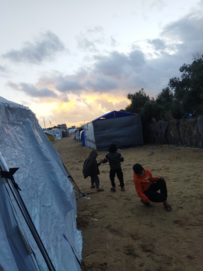 The image depicts an outdoor scene within an informal encampment in Khan Yunis, Palestinian Territory, captured during dusk or dawn under an overcast sky featuring a horizontal band of yellow-orange light at the horizon. The ground consists of unpaved sand. In the immediate foreground on the left, a large tent constructed from light-colored tarp is partially visible, showing its support poles and guy ropes. A dirt path extends into the midground, flanked by a series of makeshift structures built from tarps and assorted materials. One prominent structure in the midground-right exhibits a dark gray body and a dark blue tarp roof. To the far right, a dense line of green foliage, consistent with trees or bushes, is present, alongside a fence-like barrier constructed from organic materials. Distant buildings and utility poles with overhead lines are discernible in the background. Three individuals, consistent with children, are situated on the sandy ground in the midground. Two children on the left are standing and holding hands, both facing away from the camera; one wears a dark hooded jacket, the other a dark sweater and pants. A third child on the right, wearing an orange long-sleeved shirt and dark pants, is crouched low, facing downwards towards the sand.