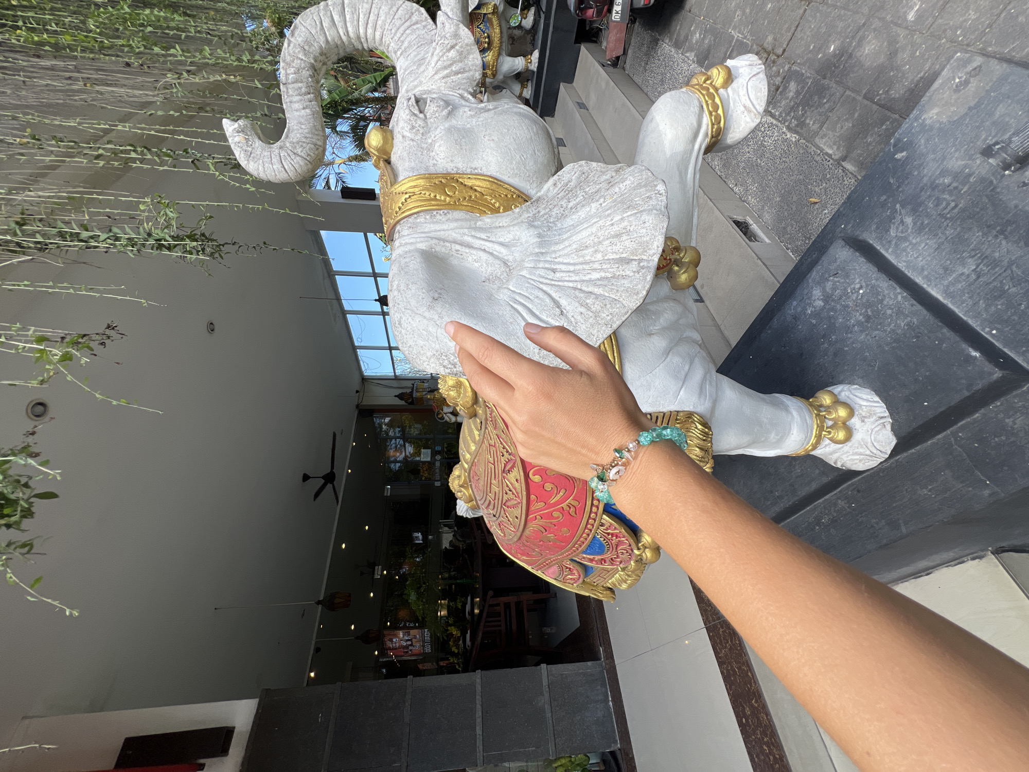 The image captures a person touching a white elephant statue in the Ubud District, Indonesia. The statue is adorned with gold accents and colorful decorative elements. The scene is indoors, evidenced by a ceiling fan, hanging lights, plants, and tables visible in the background. Through windows, one can glimpse the outside environment. A hand with a beaded bracelet is positioned on the statue. Potted plants hang from the top of the building. Stairs with a motorbike are visible in the upper part of the shot. The location is urban, with stone pavements and steps surrounding the statue.