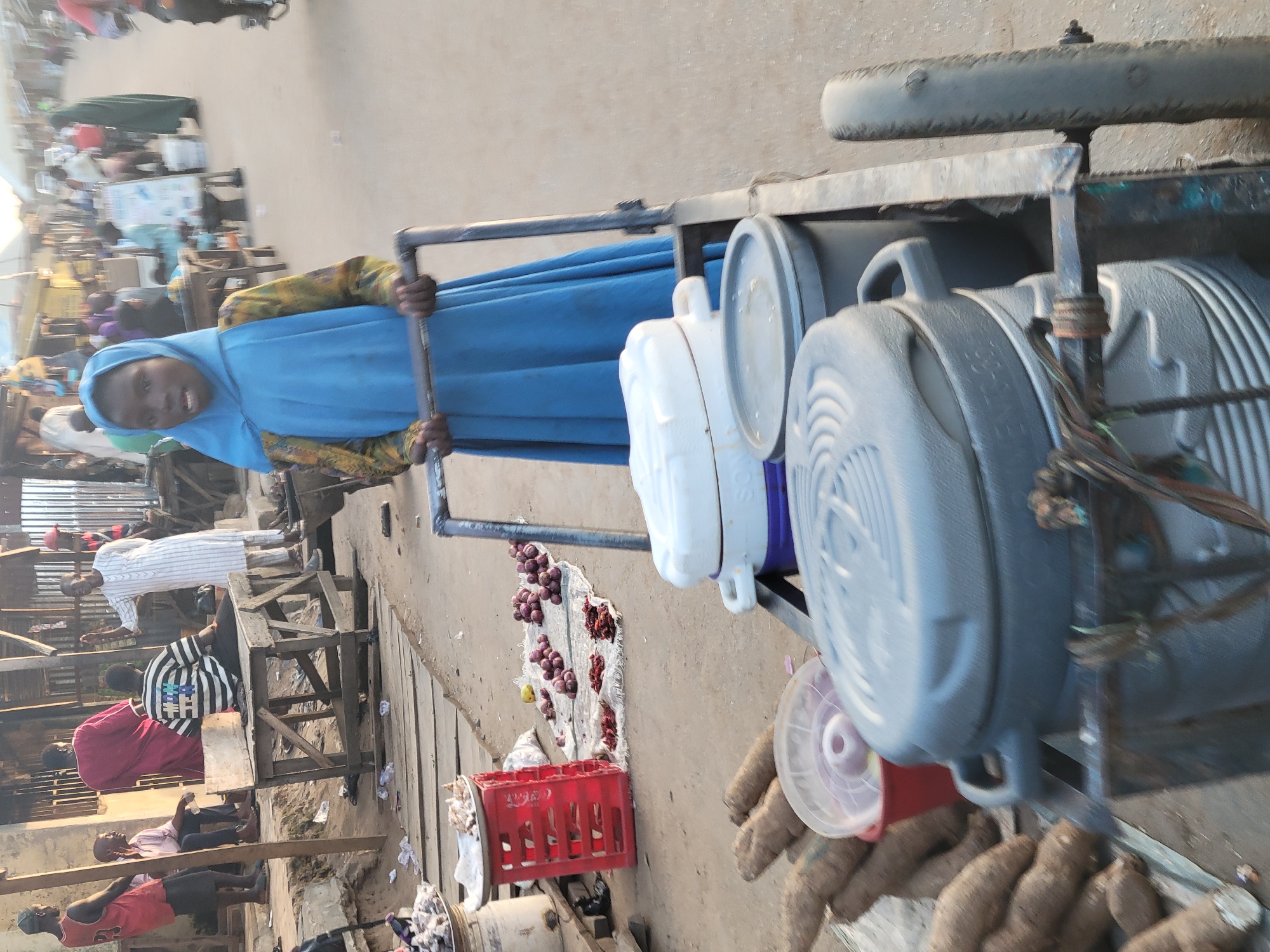 In Madalla, Nigeria, a young girl wearing a blue hijab pushes a cart with two large, gray water containers and a white container. On the cart's bed are red onions, a red crate, and yams. In the background, people are seen sitting and standing near wooden structures, suggesting a market or roadside setting. The ground appears to be a paved surface with some litter.