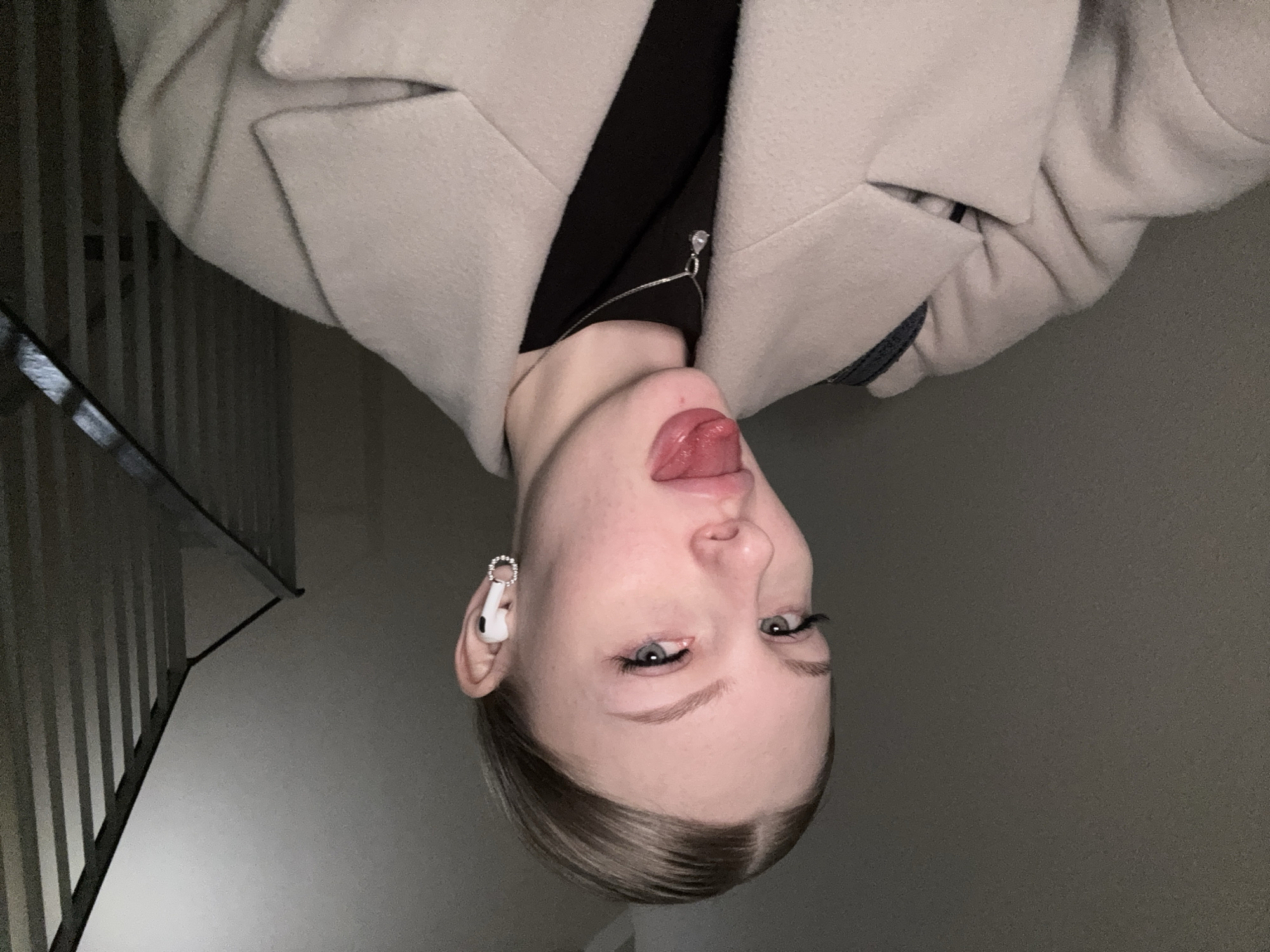 A photograph depicts a woman with pale skin and blonde hair, positioned upside down. She is wearing a light-colored coat over a black top, adorned with a delicate silver necklace. She is sticking her tongue out. An earbud is visible in her left ear, embellished with a sparkling ring. The background suggests an interior space with a staircase featuring metal railings. The location is unspecified, but the prompt mentions the city of Perm, Russia.