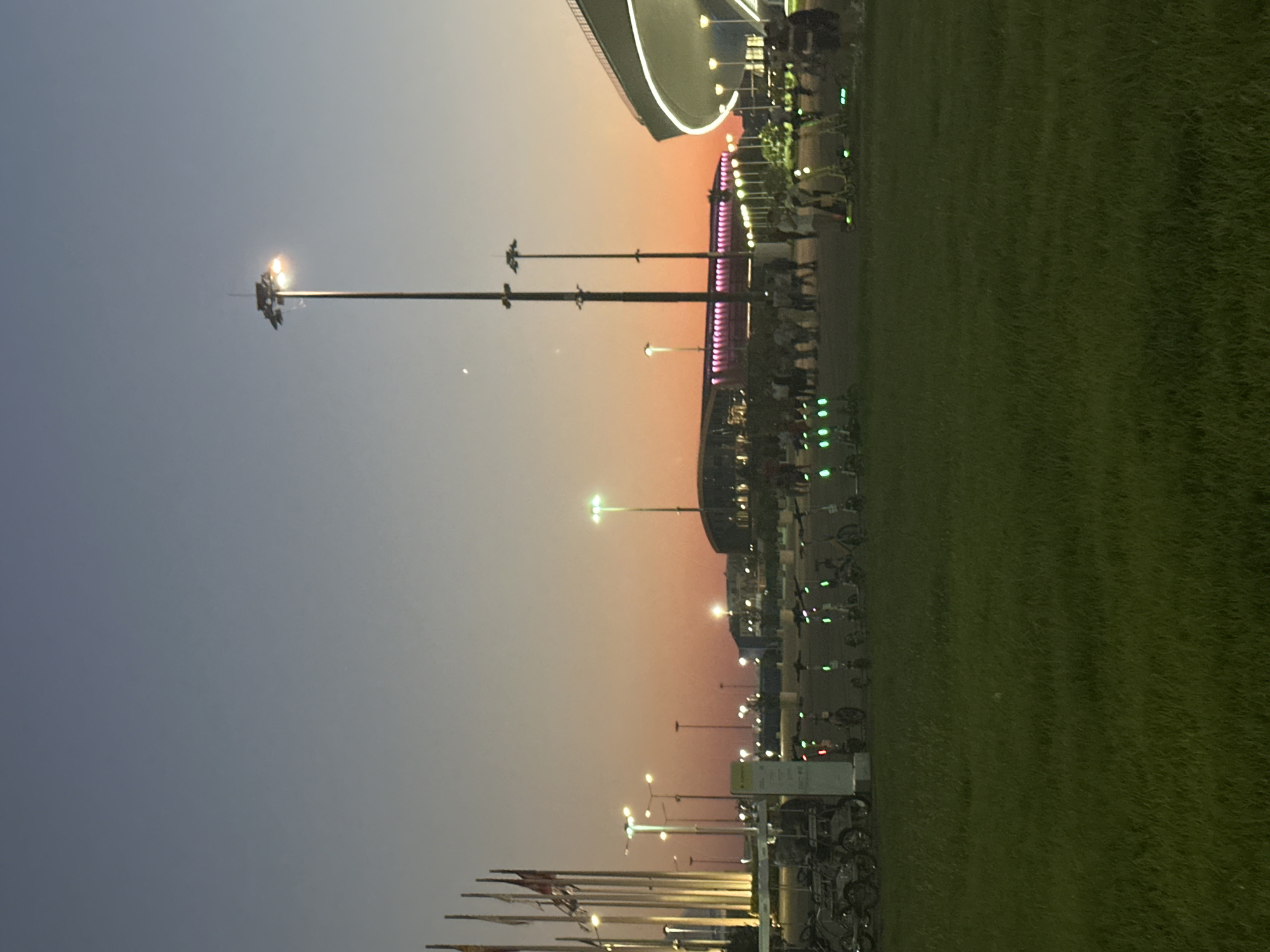 The photograph depicts an outdoor scene at dusk in Sochi, Russia, likely within the Olympic Park area, given the presence of modern architecture and an open, landscaped environment. The sky transitions from a deep blue at the top to a soft orange and pink hue near the horizon, indicating the setting sun.
Dominating the mid-ground is a row of parked electric scooters, many with illuminated green lights, suggesting they are available for rent. To the left, a collection of bicycles is also visible, parked in a designated area. Behind the scooters, a crowd of people is gathered, some standing and conversing, while others appear to be in motion.
In the background, several large, modern buildings are discernible. One prominent structure on the right features a distinctive curved roofline with bright white lighting accentuating its edge. Another building to the left displays a linear arrangement of pink lights along its facade. Numerous tall lampposts with bright lights are scattered throughout the scene, illuminating the area despite the fading daylight. Flags on tall poles are also visible on the far left. The foreground is a grassy field, contributing to the park-like atmosphere.