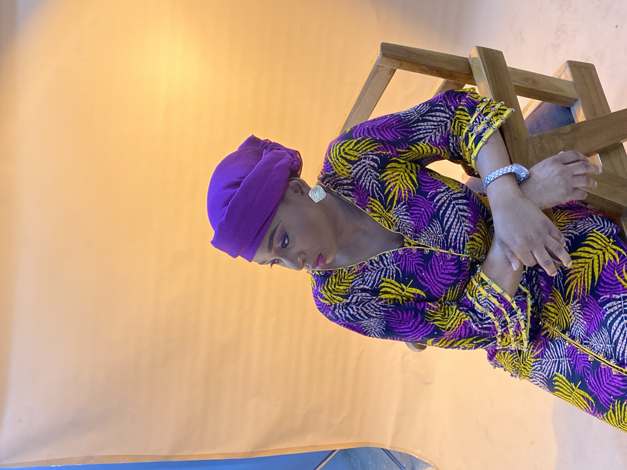 A woman in a purple headwrap and a long-sleeved top with a purple, yellow, and black palm leaf pattern is posed next to a light-colored wooden chair. She wears a silver bracelet and silver stud earrings. She is looking downward. The background is a light-yellow painted wall, and the floor is blue. The image may have been taken in a studio or a home in Gura Top, Nigeria, but specific location details are not discernible from the image.