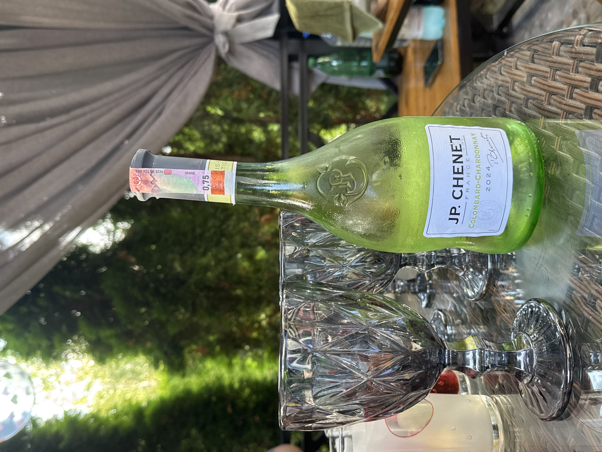 The image shows a close-up view of a table on an outdoor patio, likely in Sary-Oy, Kyrgyzstan, given the context. In the foreground, there is a chilled, pale green bottle of JP. Chenet Colombard-Chardonnay wine, dated 2024. The bottle is covered in condensation. To the left of the bottle are three grey, faceted wine glasses. The table surface is made of woven material. In the background, there are lush green trees and foliage, suggesting a natural, possibly garden setting. A grey canopy or awning is visible overhead, along with some draped fabric. Other objects, including another green bottle and a light-colored towel, are partially visible in the background, suggesting other items on the table or nearby. The lighting indicates it is daytime, with sunlight filtering through the trees.