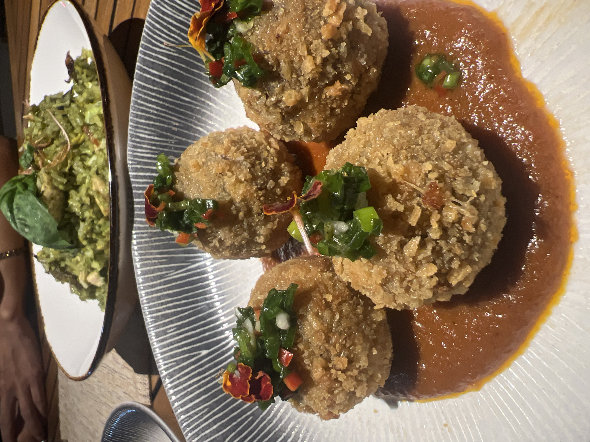 The image shows a plate of food on a table. The main dish consists of four breaded, spherical appetizers, each garnished with a mixture of chopped green vegetables and small orange flowers. These appetizers are arranged on a light brown sauce. To the left is a plate of green salad, with a large basil leaf on top. The plates are on a wooden table. A small portion of a person's arm and wrist are visible in the lower left of the image. Based on the style of food presentation and likely clientele, this could be a restaurant in the North Kuta area of Bali, Indonesia, but location verification beyond general area is not feasible based on the image alone.