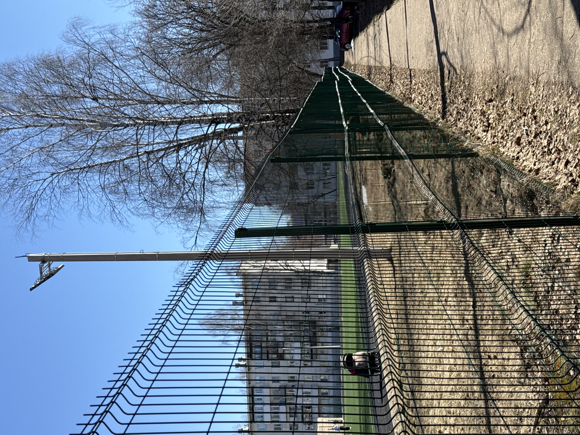The image depicts a section of wire mesh fencing in Chaikovsky, Russia, bordering a pedestrian path. The fence, constructed with horizontal and vertical wire strands, is positioned between the path and a green area. The background includes a multi-story building, likely an apartment complex, partially obscured by the fence. Above the fence, bare tree branches extend towards a clear blue sky. The path next to the fence is lined with fallen leaves and shadowed by the trees. At the top right of the scene, the rear of a car is just visible, parked along the path.