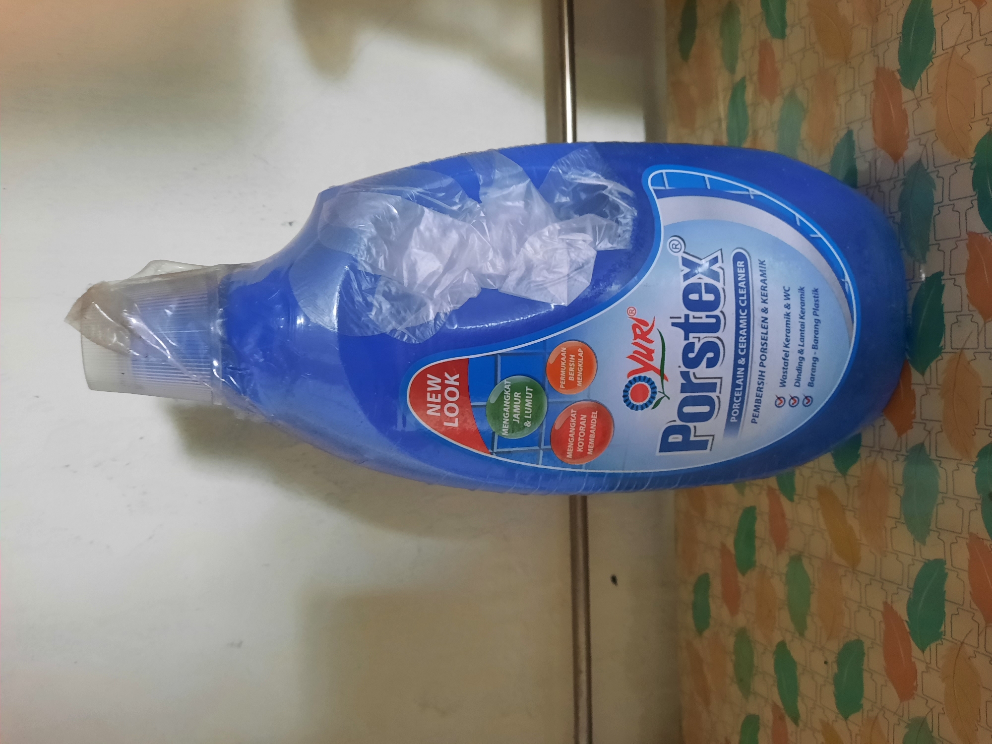 A blue bottle of "Yuri Porstex Porcelain & Ceramic Cleaner" is displayed in an indoor setting. The bottle is partially covered in transparent plastic packaging. The label on the bottle is primarily blue and white, with red and green accents. Text on the label indicates "NEW LOOK," "MENGANGKAT JAMUR & LUMUT" (Removes mold & slime), "MENGANGKAT KOTORAN MEMBANDEL" (Removes stubborn dirt), and "PERMUKAAN BERSIH MENGKILAP" (Clean shiny surface). It also states "PORCELAIN & CERAMIC CLEANER" and "PEMBERSIH PORSELEN & KERAMIK." Below this, there are bullet points: "Wastafel Keramik & WC" (Ceramic sink & WC), "Dinding & Lantai Keramik" (Ceramic walls & floor), and "Barang - Barang Plastik" (Plastic items). The bottle is placed on a patterned surface with leaf-like designs in various colors, including green, orange, and yellow, against a light-colored wall. The scene appears to be within a dwelling or shop in Lhokseumawe, Indonesia.