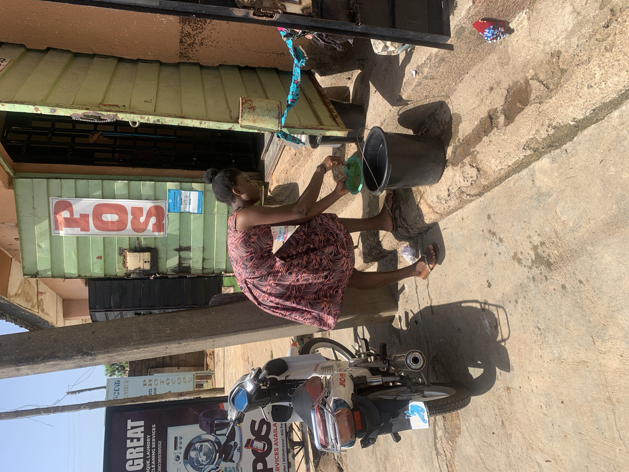 This media file, captured in Jos, Nigeria, depicts a young woman engaged in a daily chore outdoors under bright sunlight. She is seen squatting or sitting on a concrete ground, wearing a patterned dress, and is focused on washing something in a green plastic bowl. Two black buckets are placed nearby, likely holding water for her activity.
The scene is an urban or semi-urban setting, possibly an alleyway or a side street. Behind the woman, there's a building or stall with a light green corrugated metal roof and a dark structure below it. A white and red JINCHENG brand motorcycle is parked to the left, indicating vehicle presence. The concrete ground is worn and shows patches of moisture.
Visible text provides commercial cues: a sign on the far left advertises "GREAT UNIQUE, LAUNDRY CLEANING SERVICES" with a phone number "08036938552". Another sign, possibly rotated, prominently displays "POS" (Point of Sale), corroborated by text on the motorcycle saying "POS SERVICES AVAILA...sfer | Withdrawal | Bill Pay". A blue sign adds "PAY WITH TRANSFER 312229 1643". The strong shadows suggest a clear, sunny day, likely morning or afternoon. The image captures a routine moment of life in a Nigerian city.