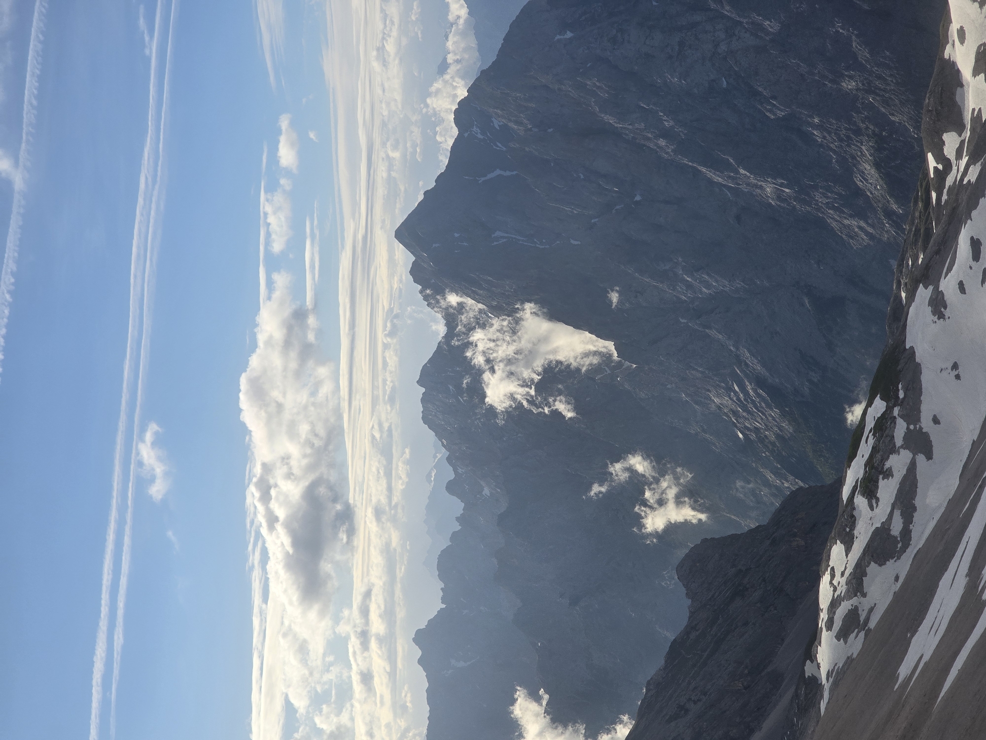 The image depicts a panoramic view of a rugged mountain range under a partly cloudy sky. In the foreground, a slope is partially covered with snow and patches of dark soil or rock. Further back, numerous jagged mountain peaks rise, some with snow-capped summits. Wisps of clouds cling to the mountainsides, adding a dramatic effect. The sky is a bright blue with scattered white cumulus clouds and long, thin contrails from aircraft. The lighting suggests it is daytime, likely morning or afternoon, with the sun illuminating the peaks and casting shadows. The scene is devoid of any people, structures, or immediate signs of human activity, evoking a sense of remote, natural grandeur.
This vista is likely captured from a high vantage point in or around Garmisch-Partenkirchen, Germany, a region known for its dramatic Alpine scenery, including the Zugspitze, Germany's highest peak.