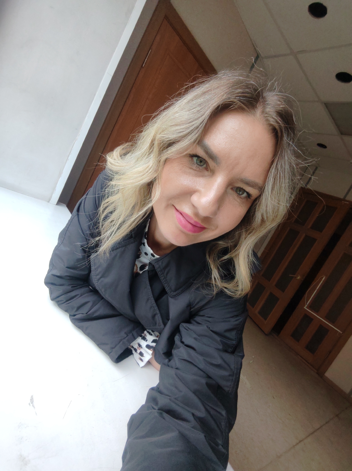 A woman with blonde, wavy hair and pink lipstick is looking directly at the camera with a slight smile. She is wearing a black, quilted jacket over a spotted blouse. The background includes a white surface she is leaning on, and behind her, there is a wooden doorframe, a dark wooden door, and a tan tiled floor. The ceiling has recessed lighting, suggesting an indoor location in a building. The location is described as Unknown, Unknown.