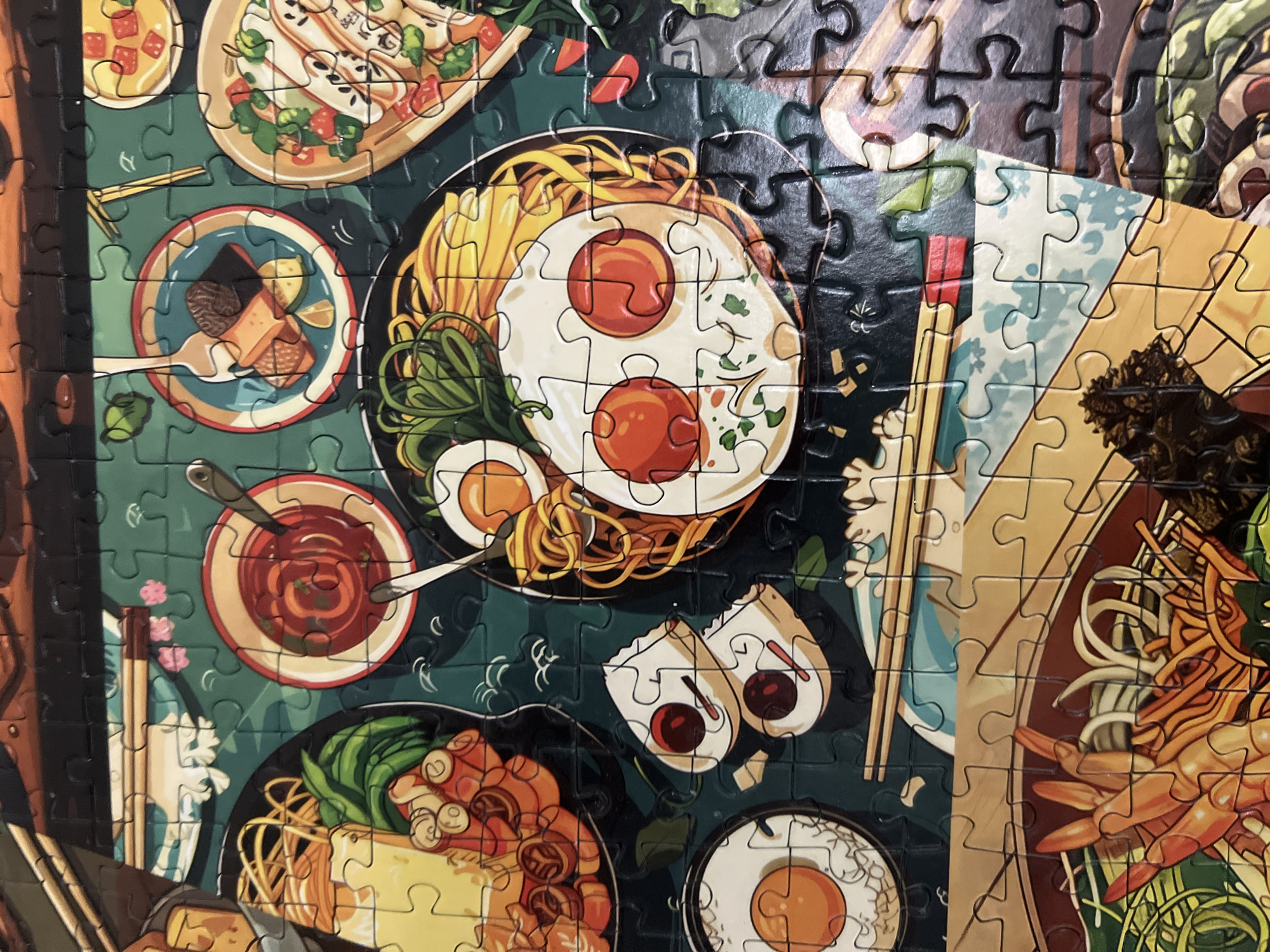 The image is a close-up, top-down view of a jigsaw puzzle depicting various Asian dishes, likely ramen and other noodle dishes, along with accompaniments. The puzzle pieces are visible, showing the distinct interlocking shapes. The food items include ramen noodles, fried eggs, shrimp, vegetables, and small bowls of sauce or broth. The background is a dark, textured surface, possibly representing a table or placemat. There are no people or animals in the image, and no discernible elements that would place it in Volgograd, Russia.