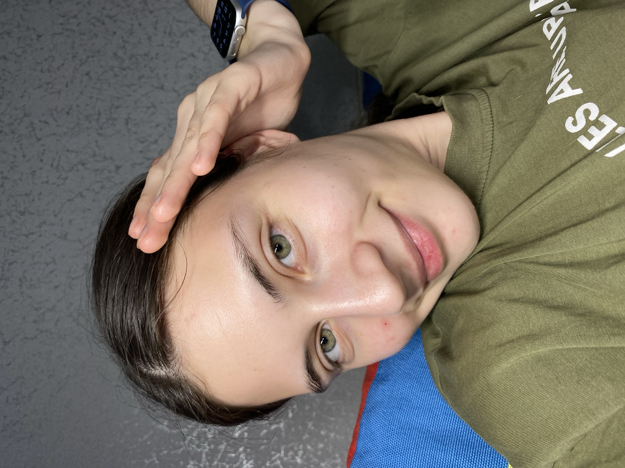 A close-up, tilted head-and-shoulders shot depicts a light-skinned person with dark hair, holding their hand to their forehead. The person's eyes are green. They are wearing a khaki-colored t-shirt with white lettering. A watch with a blue band is visible on the person's wrist. The background includes a gray surface and a blue/red patterned textile. No information indicates this photo was taken in Votkinsk, Russia.