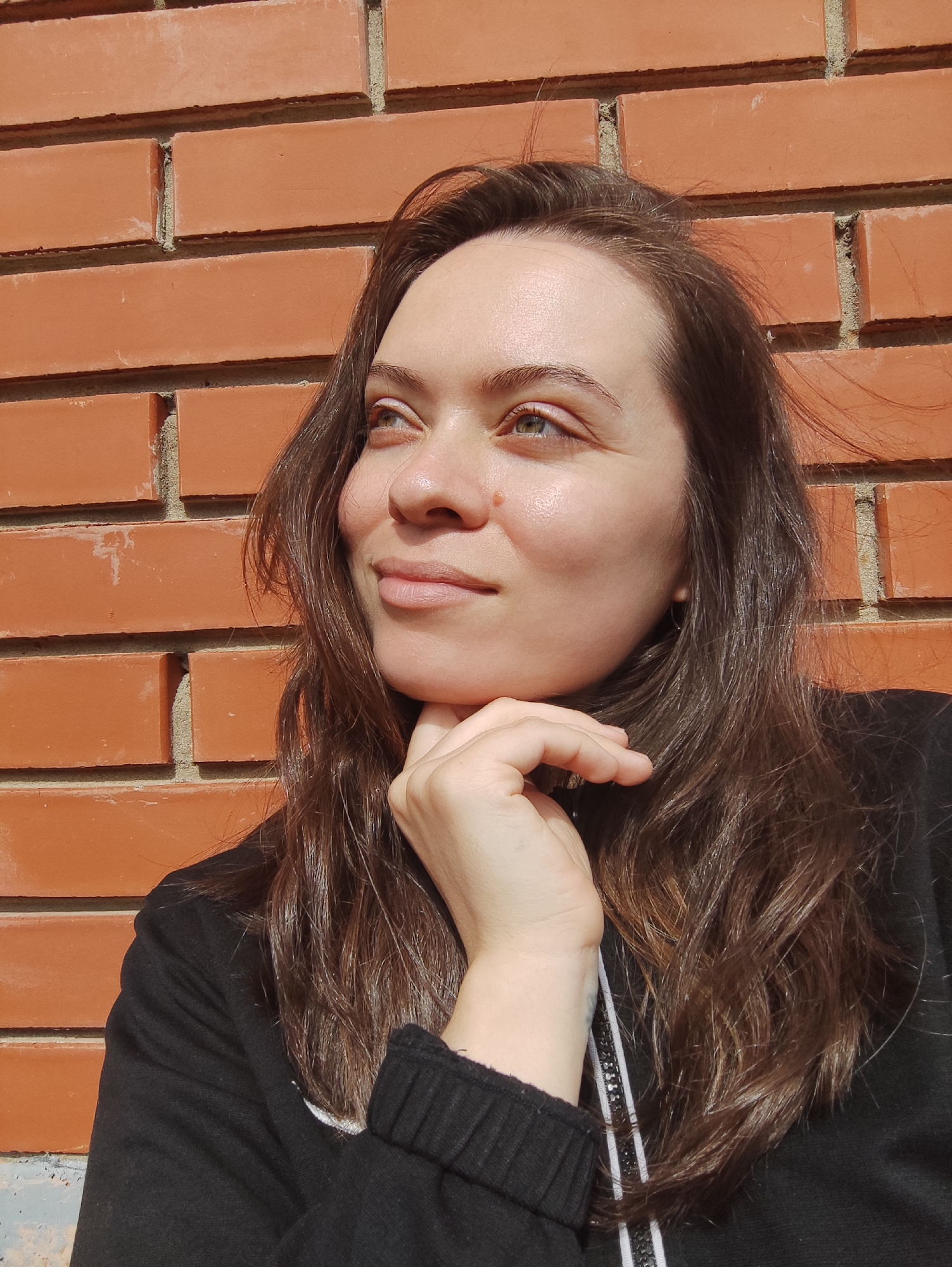 A young adult female stands close to a brick wall, her hand supporting her chin. She is dressed in a dark, possibly black, zipped top, with a white stripe visible near the zipper. She has long, dark brown hair and fair skin. The background is a red brick wall under bright lighting conditions, likely sunlight. The location is potentially Фоки, Russia.